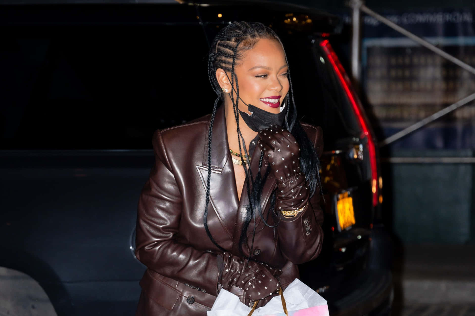 Rihanna 身穿棕色皮褸和飾釘手套，在夜晚的車旁微笑。