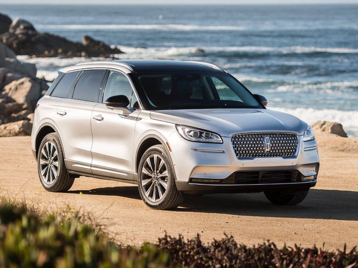 2021款林肯Corsair小型SUV，尊貴典雅 壁紙 — 一架時尚的銀色林肯SUV停泊在陽光普照的海岸公路上，背後是洶湧的海浪和岩石峭壁。