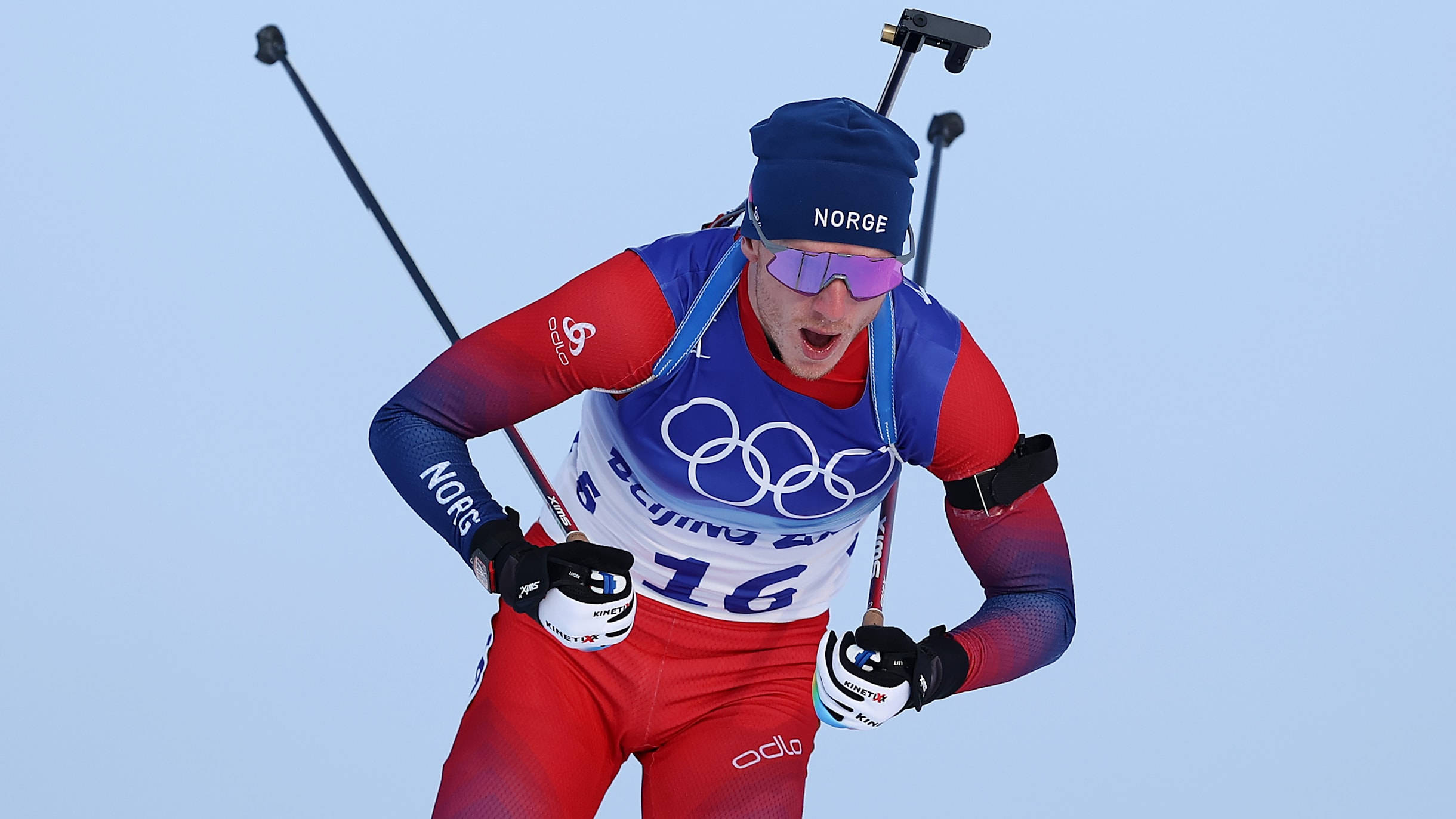 挪威越野射擊運動員約翰內斯·廷格內斯·博2022奧運會 壁紙 — 一位身穿紅藍白裝備的挪威冬季兩項運動員，在冬季奧運會比賽中，手持滑雪杖和步槍，在淡藍色天空下奮力向前。