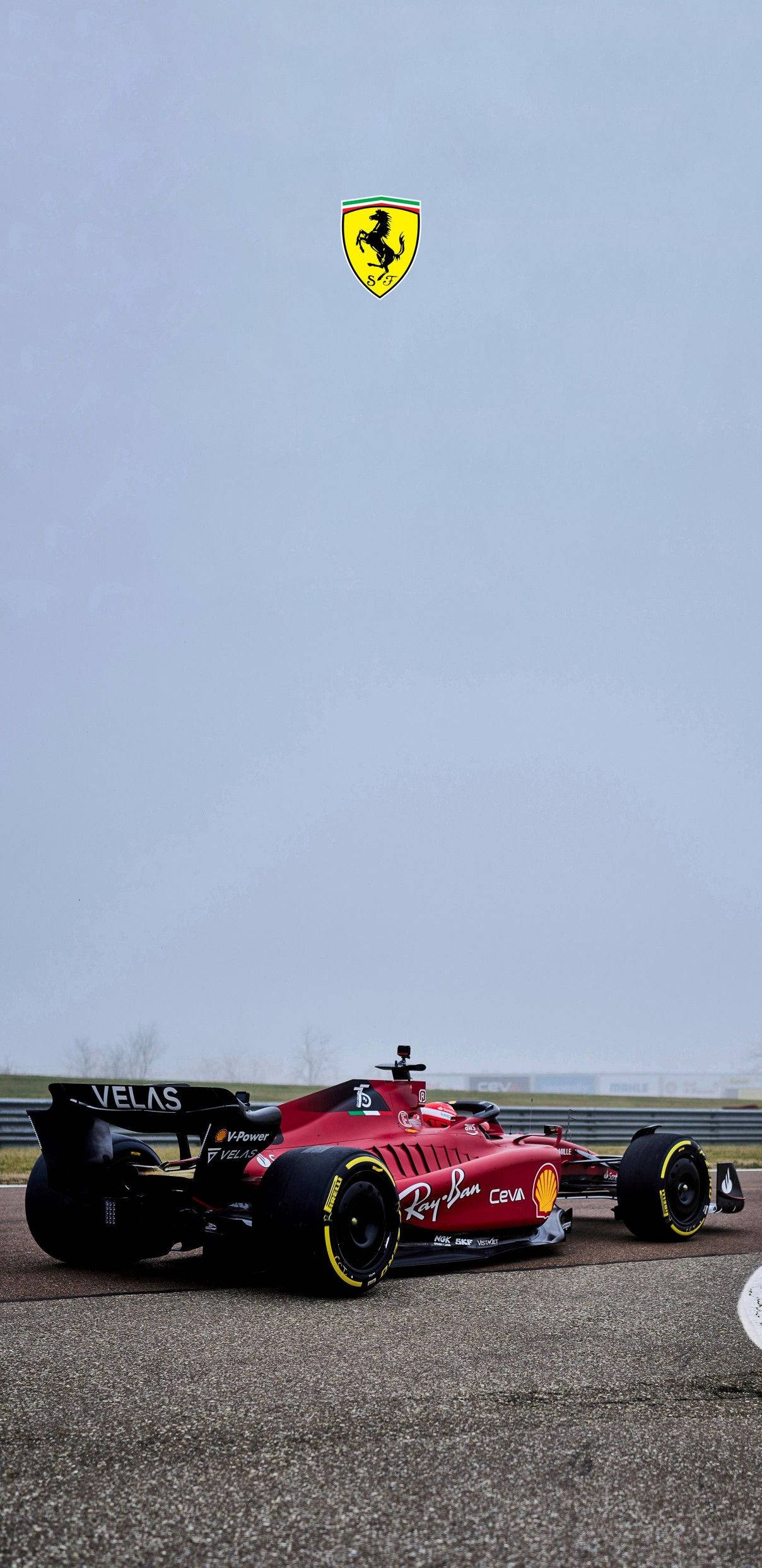 2022 F1-75法拉利手機壁紙（帶標誌） 壁紙 — 一輛紅色法拉利一級方程式賽車停在灰色天空下的賽道上，法拉利標誌漂浮在上方。