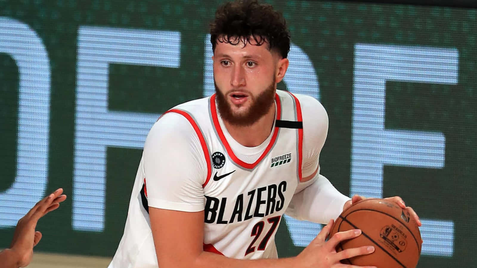 尤素福·努爾基奇2022年泡泡球場 Playoff 壁紙 — 一名專注的籃球運動員身穿白色 Blazers 球衣，在比賽中持球，背景為電子記分牌。
