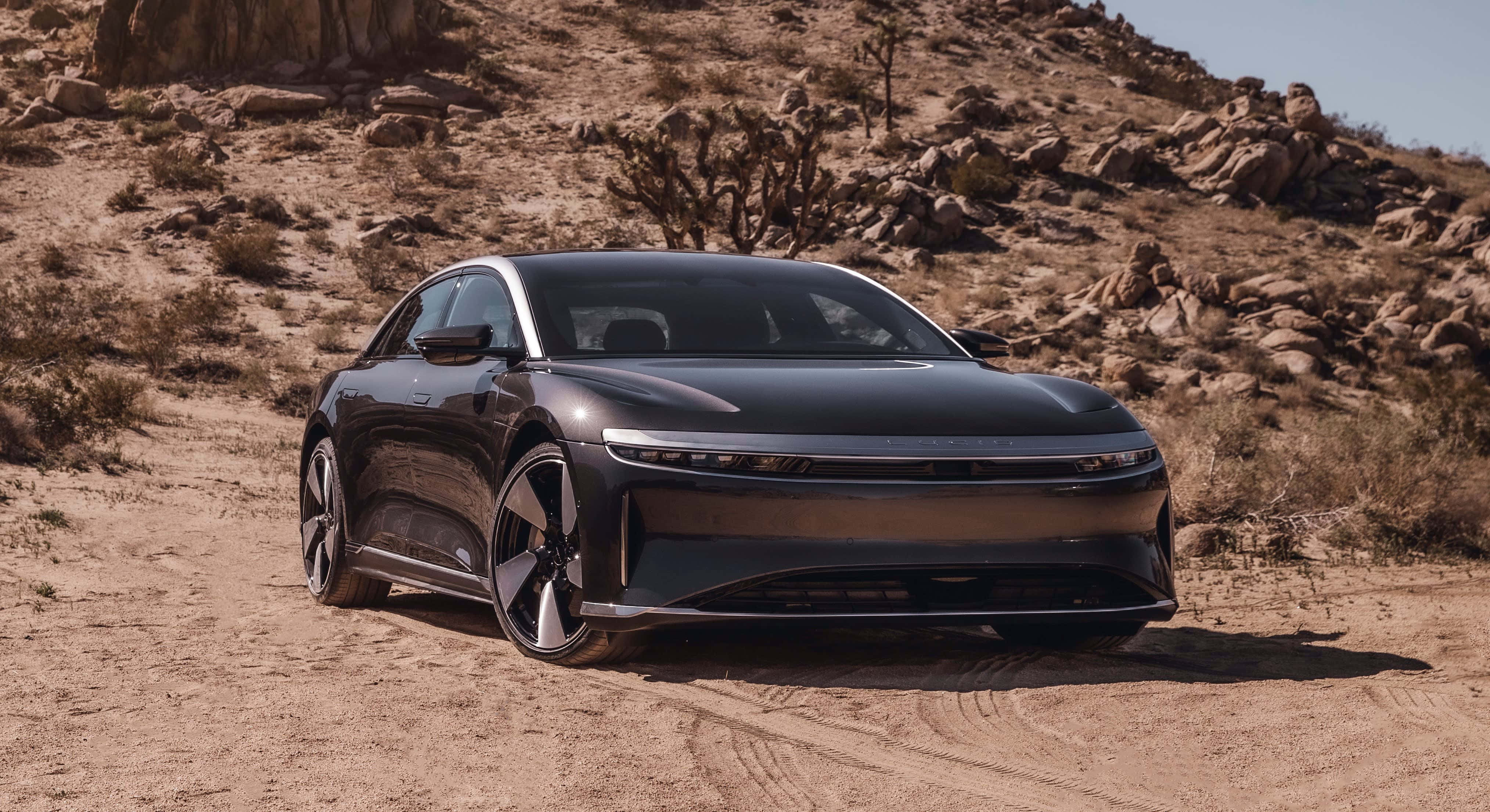 2022款Lucid豪華汽車圖片 壁紙 — 一架時尚的黑色電動車停在陽光明媚的沙漠小徑上，背景是岩石山丘和仙人掌。