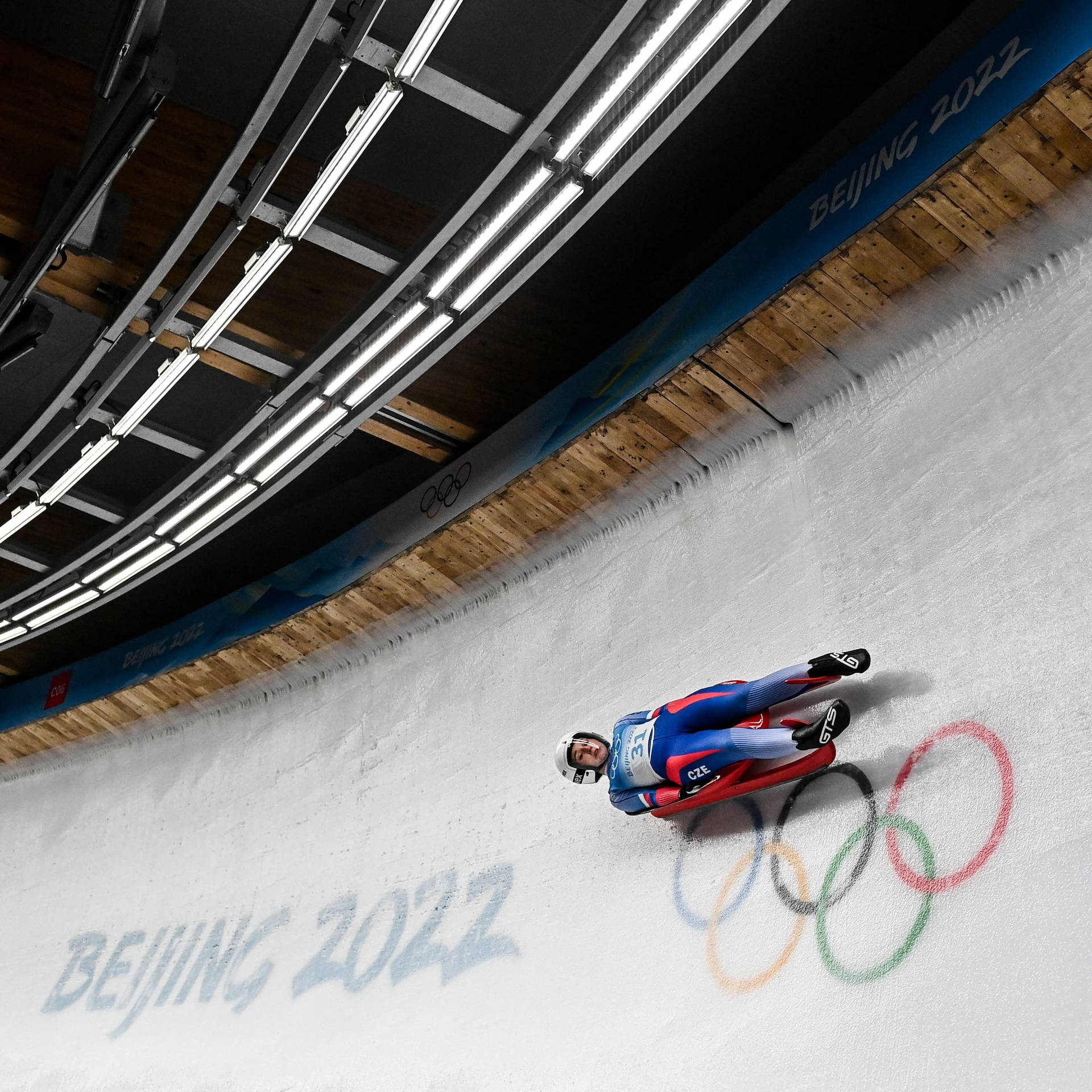 2022北京冬季奧運會美國女子單人雪橇選手 壁紙 — 一位雪橇運動員在北京2022年冬奧會的室內冰道上飛馳，冰面上繪有奧運五環。