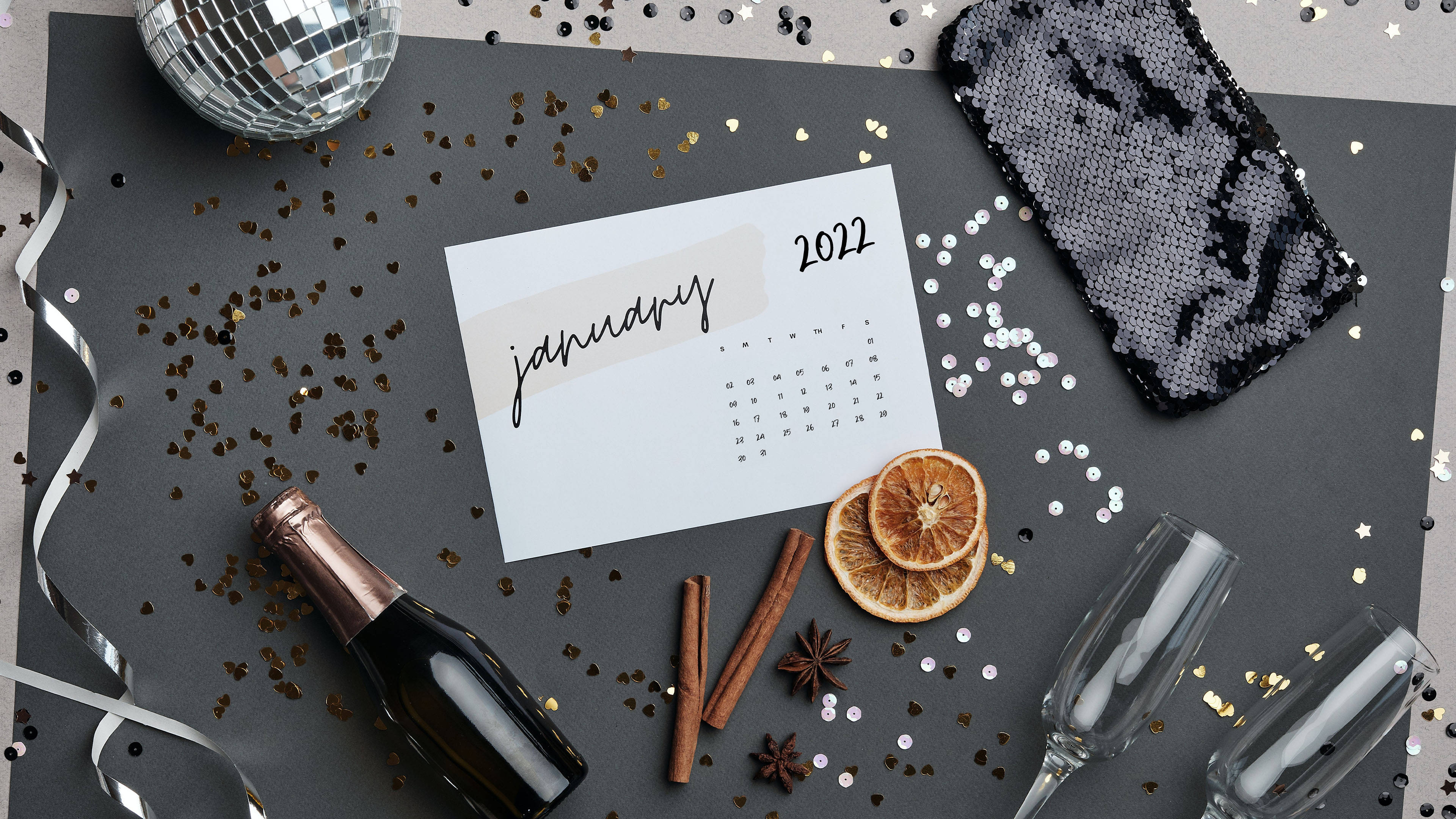 2022年1月慶祝場景 壁紙 — 一個節日平鋪構圖，包含2022年1月日曆、香檳瓶、乾柑橘、肉桂和一個迪斯科球，放置在深色表面上。
