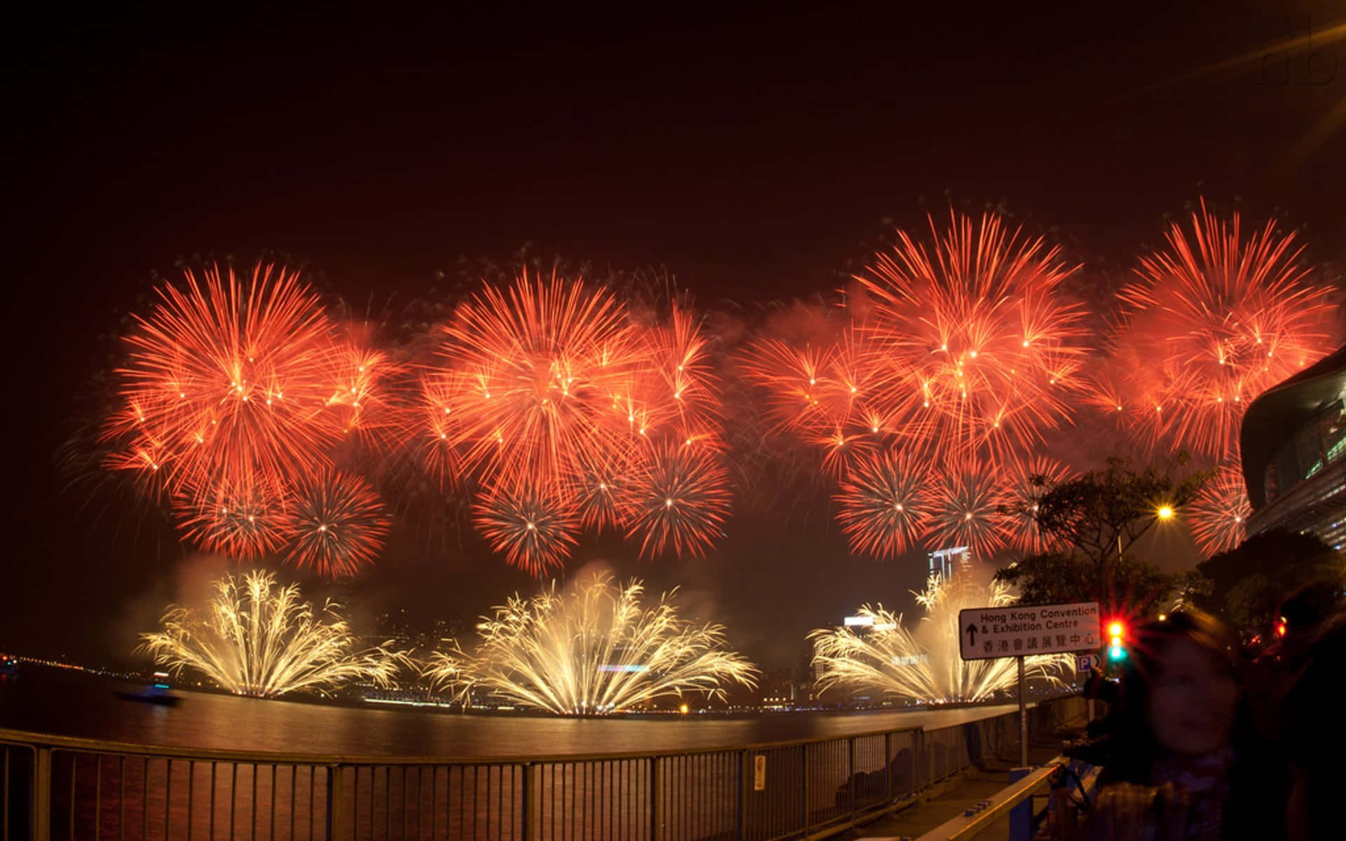 2022春節中國煙火 壁紙 — 鮮豔的紅色和金色煙火在夜間的海濱上空綻放，觀眾從欄杆旁觀看。