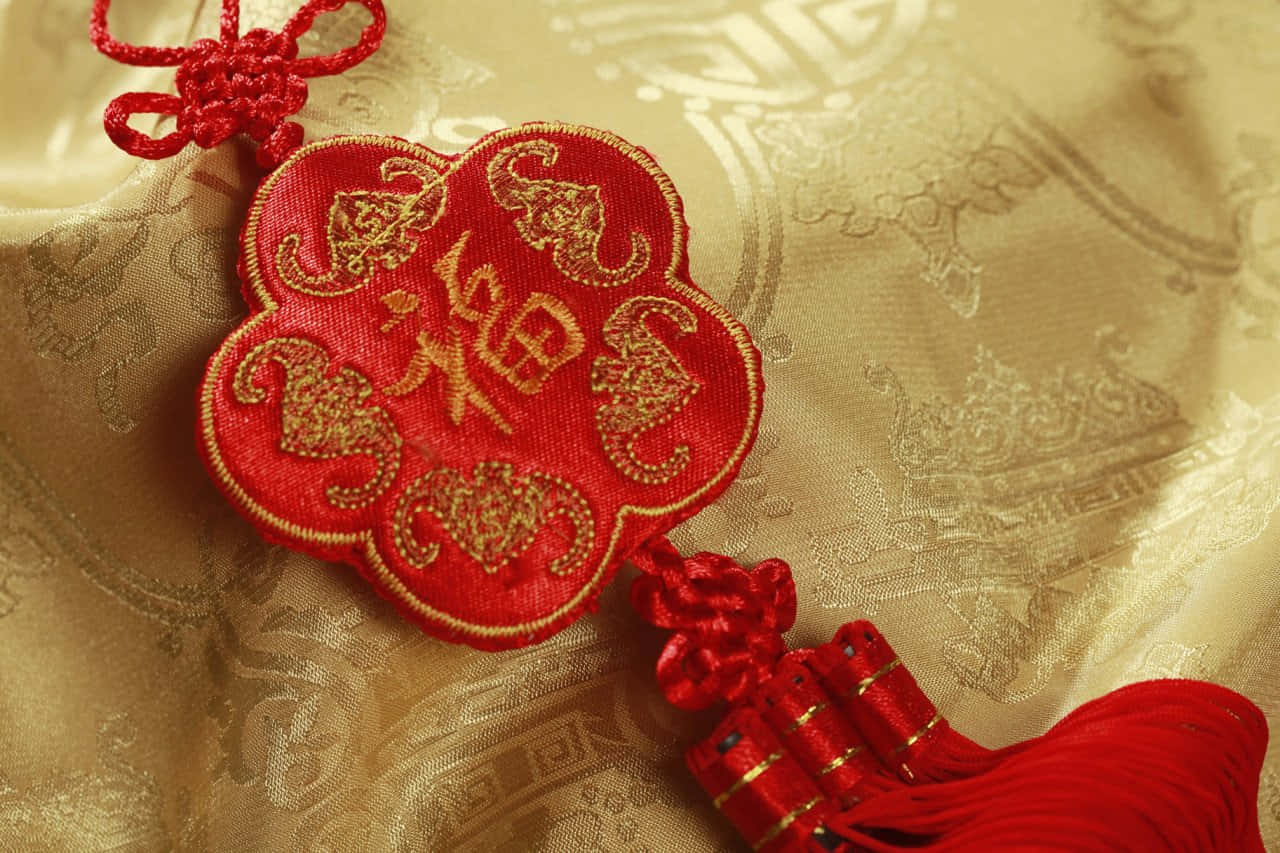 2022春節吉祥符 壁紙 — 一個鮮豔的紅色中國結飾品，帶有金色刺繡和流蘇，放置在帶有紋理的金色布料上。
