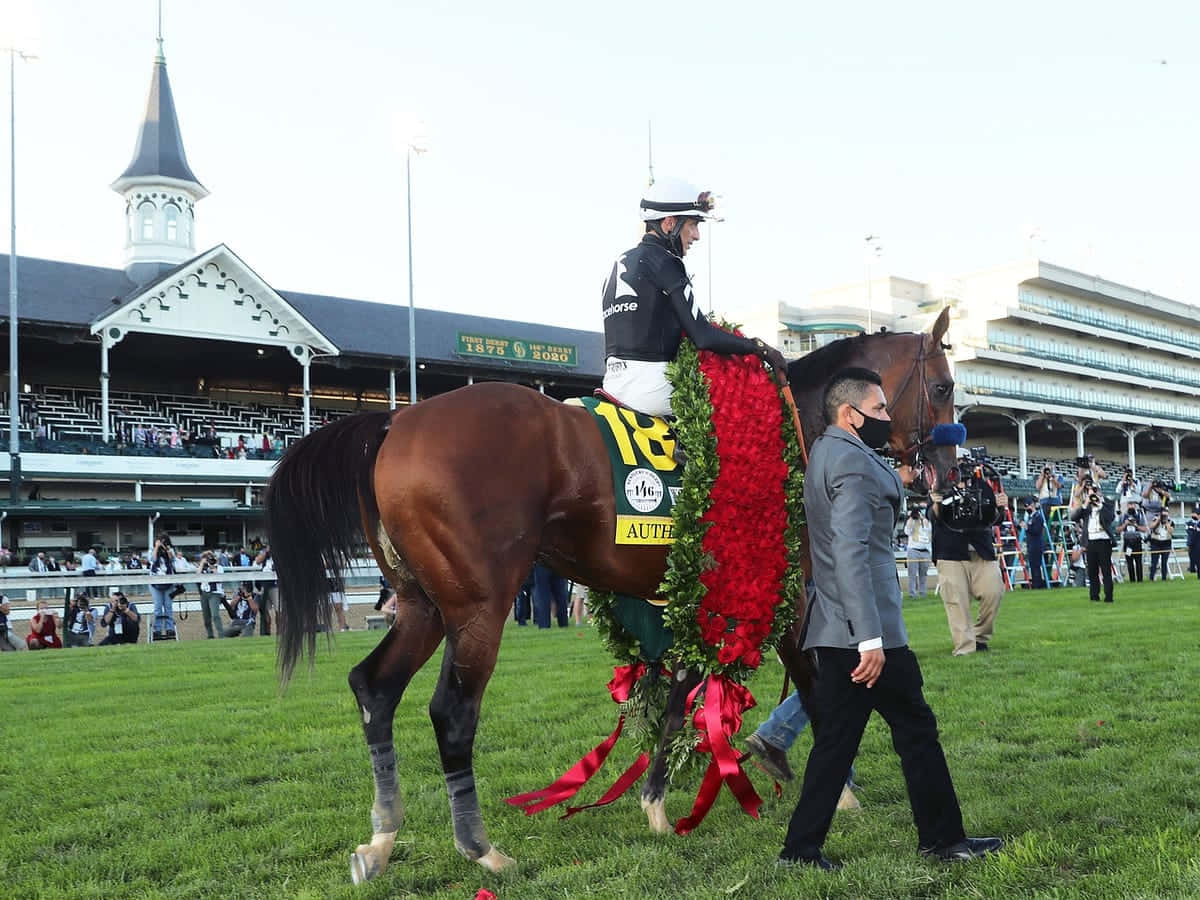 2022肯塔基德比的壯麗馬匹 壁紙 — 一匹勝利的栗色賽馬，飾有紅色花環，由騎師和馬伕帶領穿過賽馬場的草地跑道。