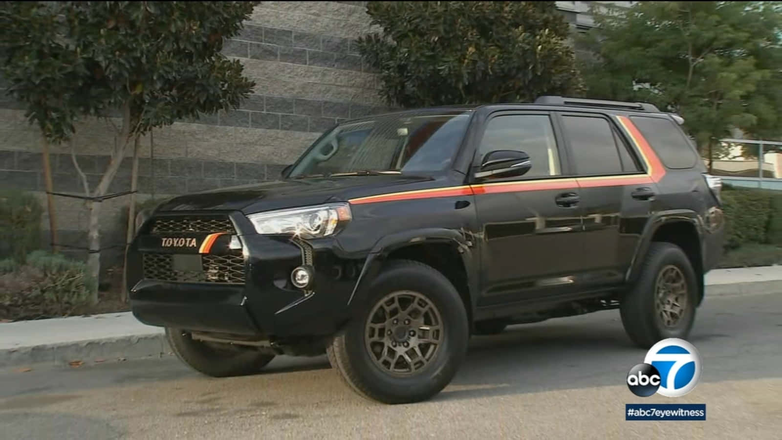 駕駛冒險，全新2023款豐田4Runner 壁紙 — 一輛帶有橙色條紋的黑色豐田4Runner SUV停放在石牆和綠化旁邊的街道上。