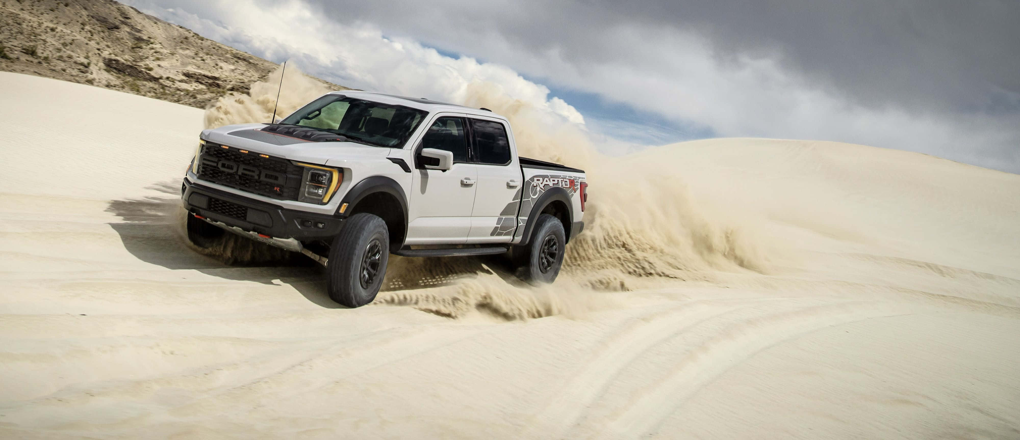 2023款福特F-150 Raptor R 壁紙 — 一架白色福特F-150 Raptor貨車在多雲的天空下駛過廣闊的沙漠沙丘，揚起沙塵。