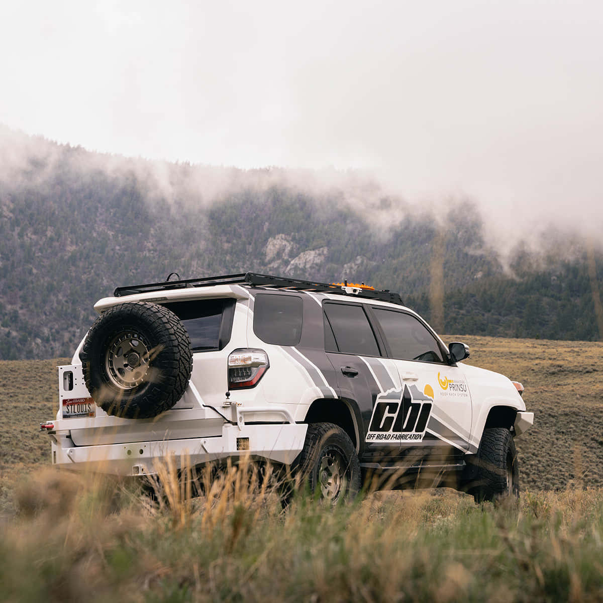 全新2023款Toyota 4Runner，造型亮眼 壁紙 — 一輛白色越野SUV，裝有備用輪胎，停放在草地上，背景是霧濛濛的山脈。