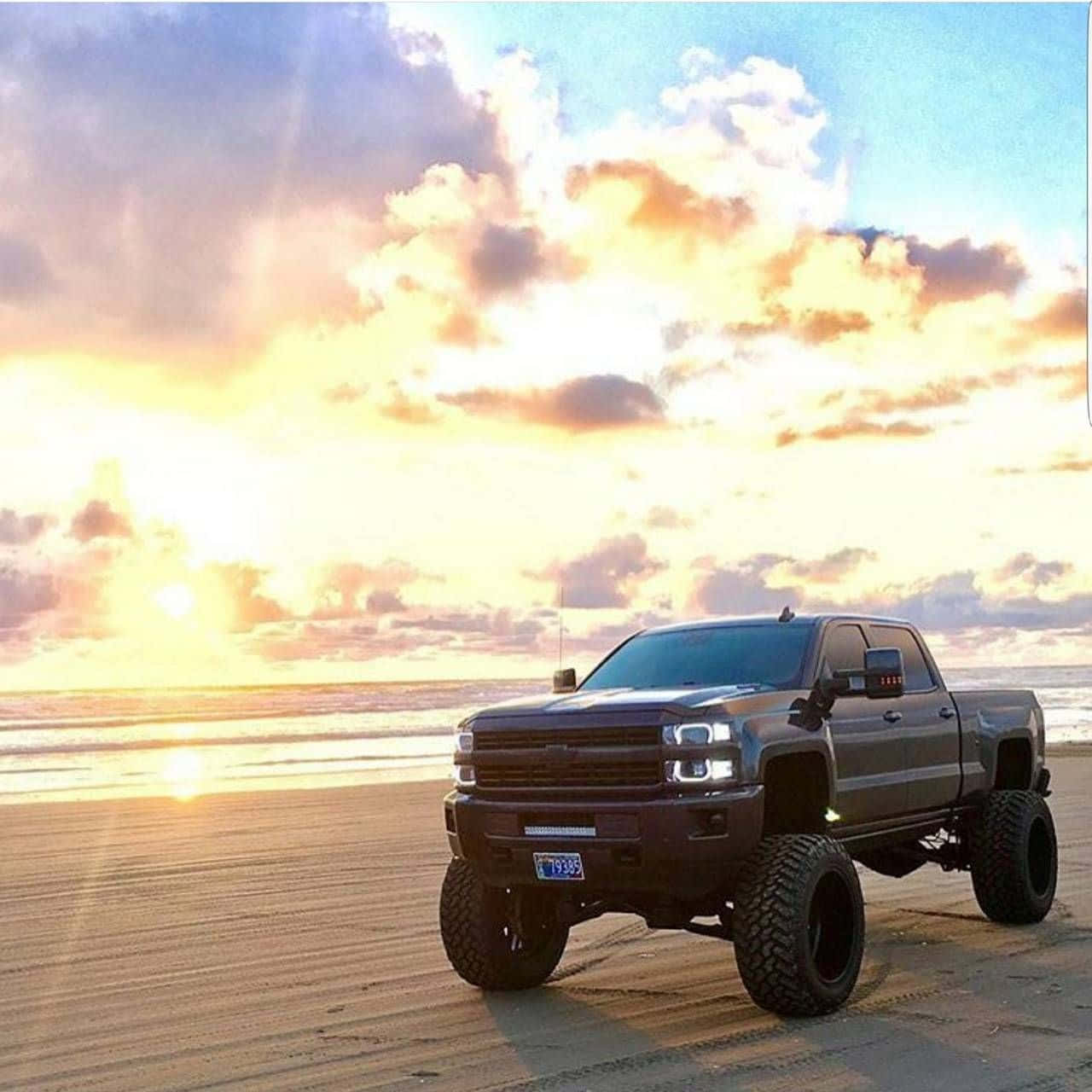 2023年黑色雪佛蘭Silverado 1500皮卡 壁紙 — 一輛升高了的黑色皮卡車停泊在沙灘上，夕陽西下，金色雲彩與海浪相伴。