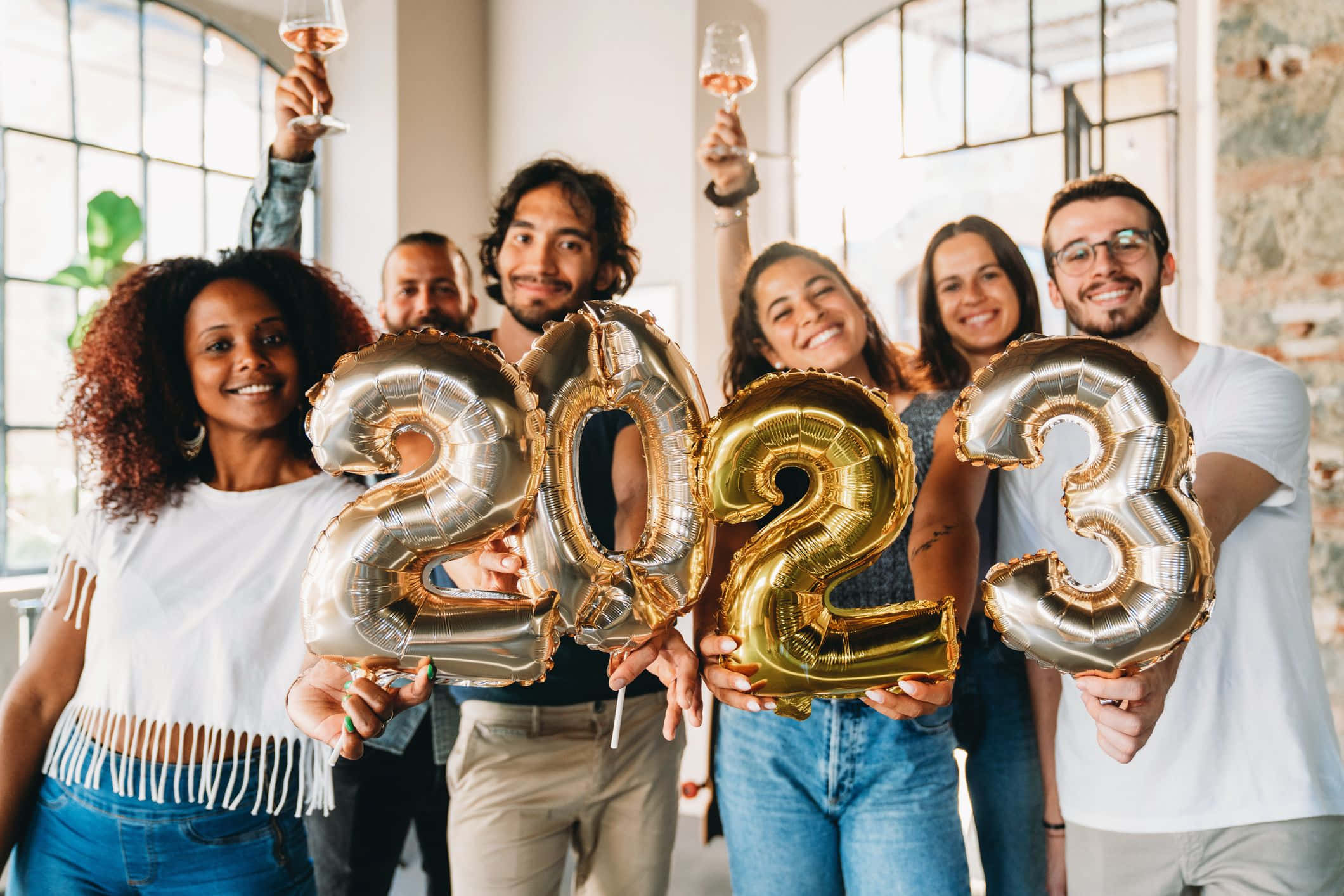 2023新年慶祝圖 壁紙 — 一群快樂的朋友在明亮的閣樓裡用金色和銀色的2023氣球和玫瑰紅酒慶祝除夕。