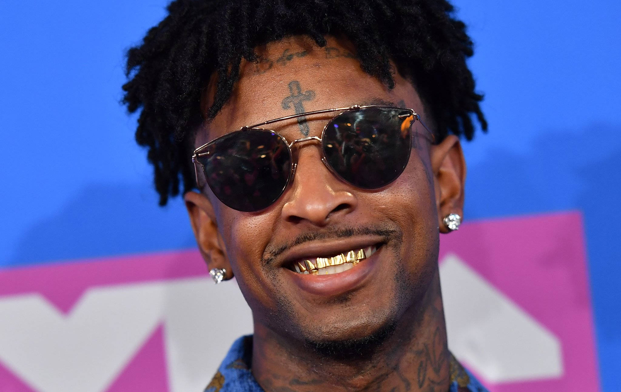 21 Savage 2018 MTV 頒獎典禮 壁紙 — 一個微笑的男人，留著辮子、金牙和太陽眼鏡，在充滿活力的藍色和粉紅色MTV背景前擺姿勢。