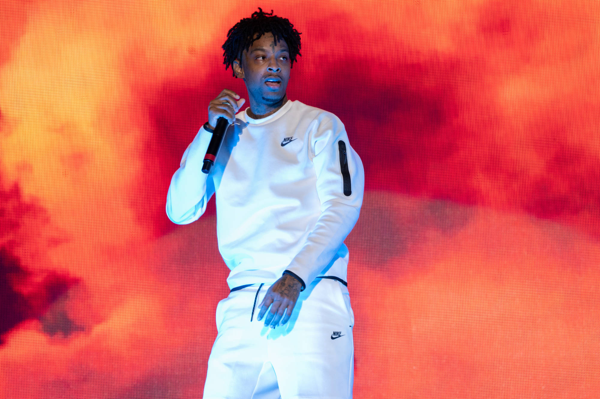 21 Savage 2021 總督音樂節 壁紙 — 一位身穿白色Nike運動服的饒舌歌手在火紅與橙色燈光下於舞台上手持麥克風表演。