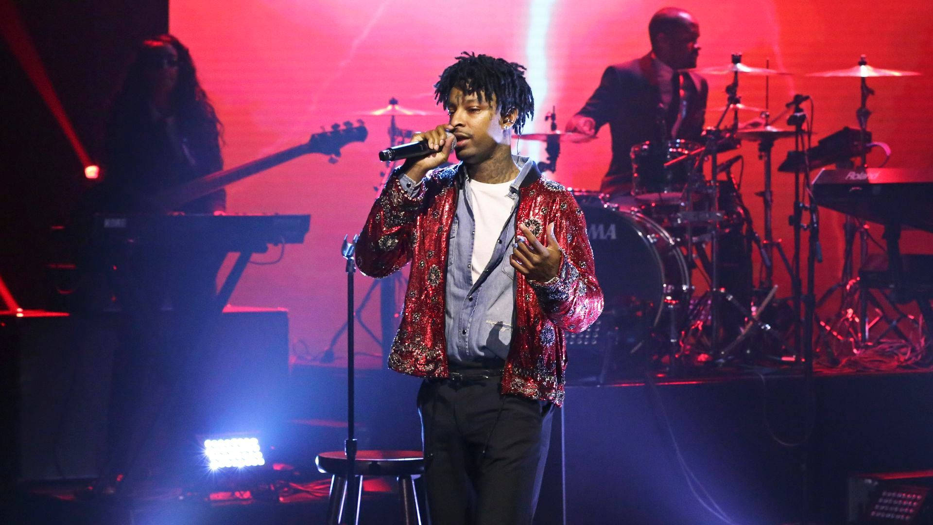 21 Savage《A Lot》現場演出 壁紙 — 一位身穿紅色圖案外套的歌手在舞台上表演，背後有樂隊，舞台上有鮮豔的紅色和藍色燈光。
