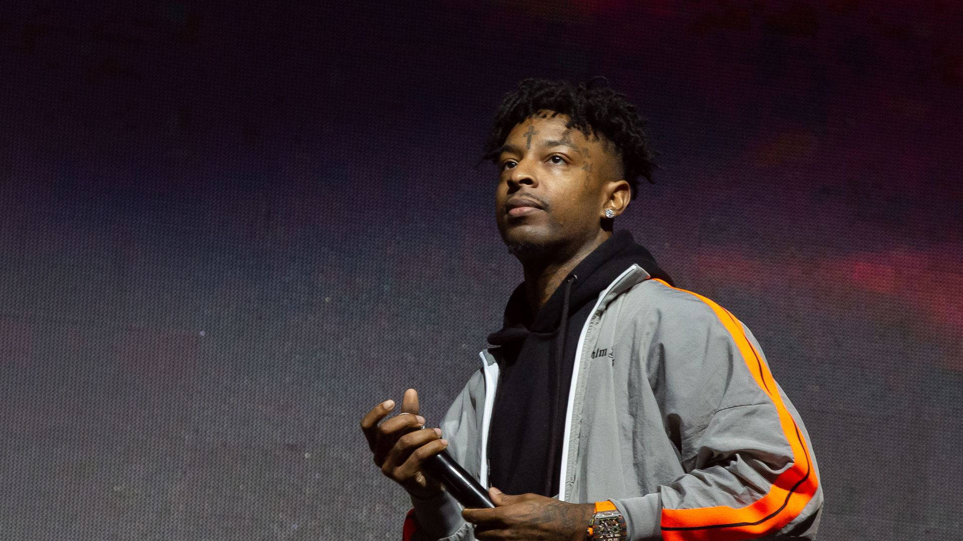 21 Savage ACL 音樂節 壁紙 — 一位男性饒舌歌手留著辮子，在戲劇性的紫色和紅色燈光下在舞台上拿著麥克風。