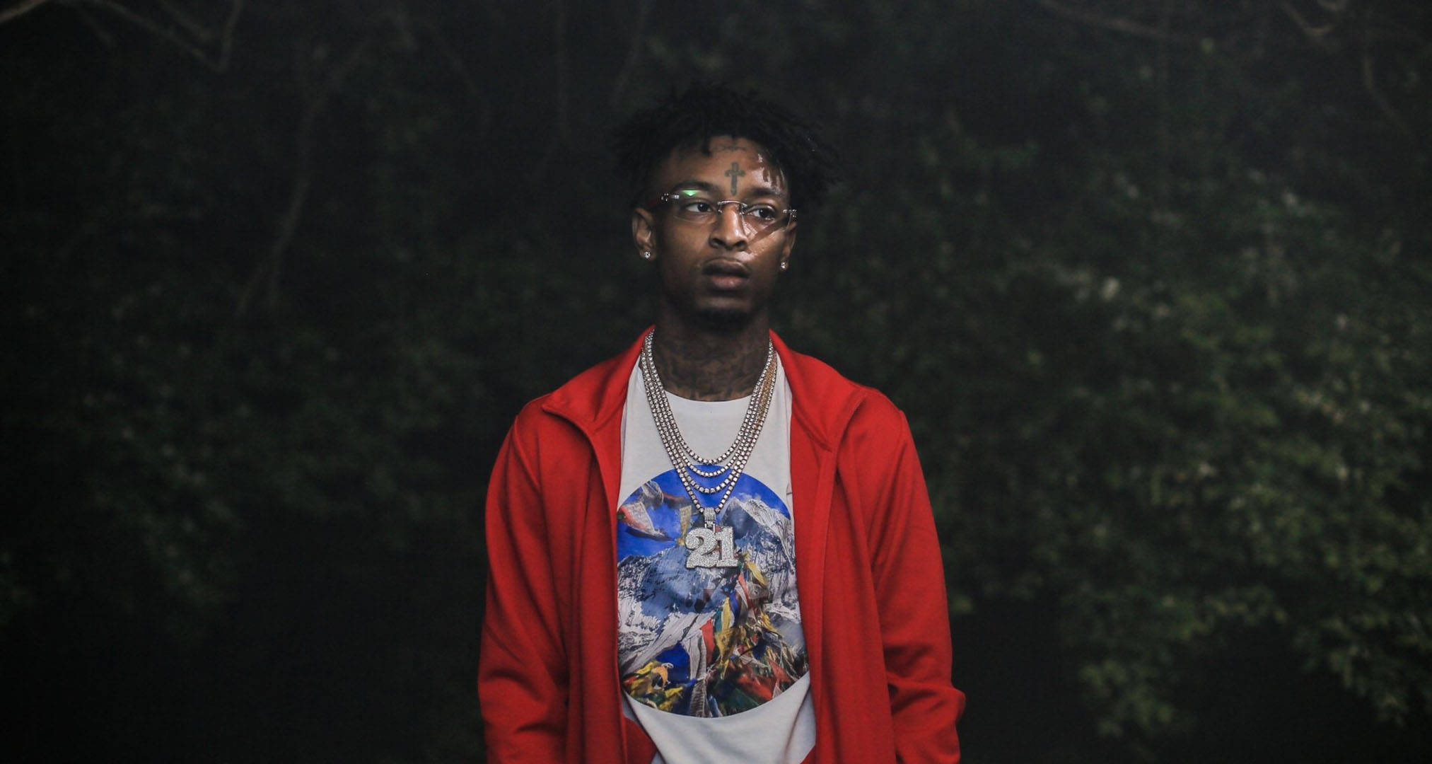 21 Savage《All The Smoke》MV截圖 壁紙 — 一名穿著紅色連帽衫和圖案T恤的年輕男子在夜晚的黑暗森林背景下自信地站立。