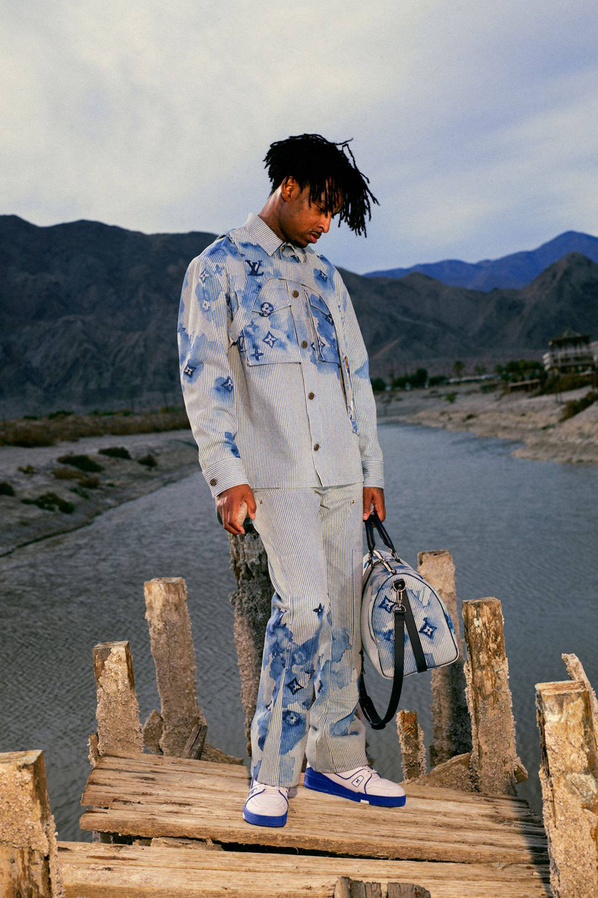 21 Savage LV 夏季服裝 壁紙 — 一名身穿藍色圖案路易威登服裝的男子站在一條風化的木橋上，橋下是河流，背景有山脈。