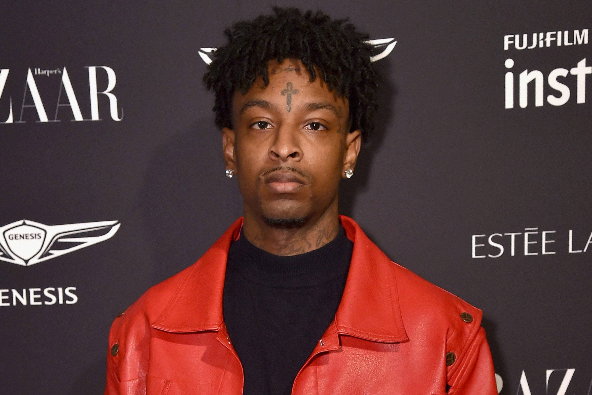 21 Savage 出席 Harper's BAZAAR 活動 壁紙 — 一個有捲髮和十字架紋身的男人，在品牌活動背景前穿著紅色皮褸，嚴肅地凝視著。