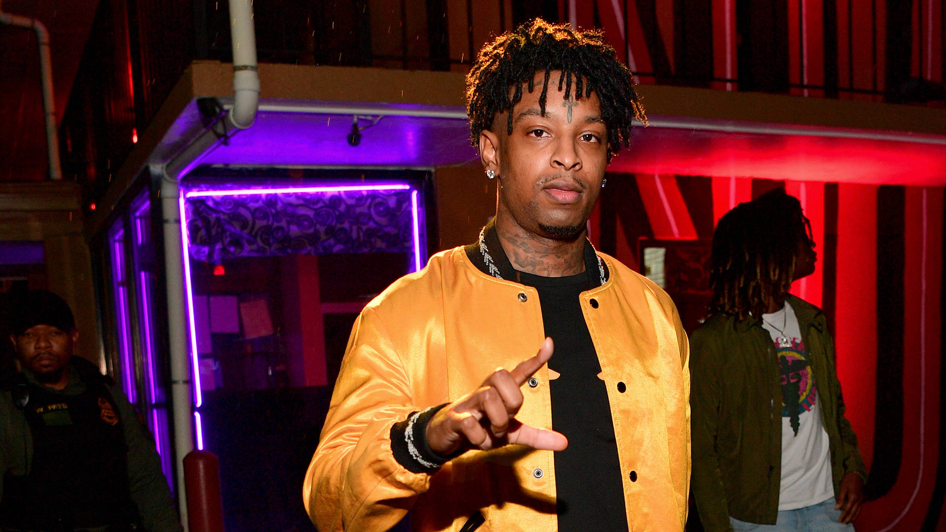 21 Savage 出席 Motel 21 壁紙 — 一名身穿閃亮黃色外套的男子在昏暗的城市小巷中，在霓虹紫和紅燈光下向前指。