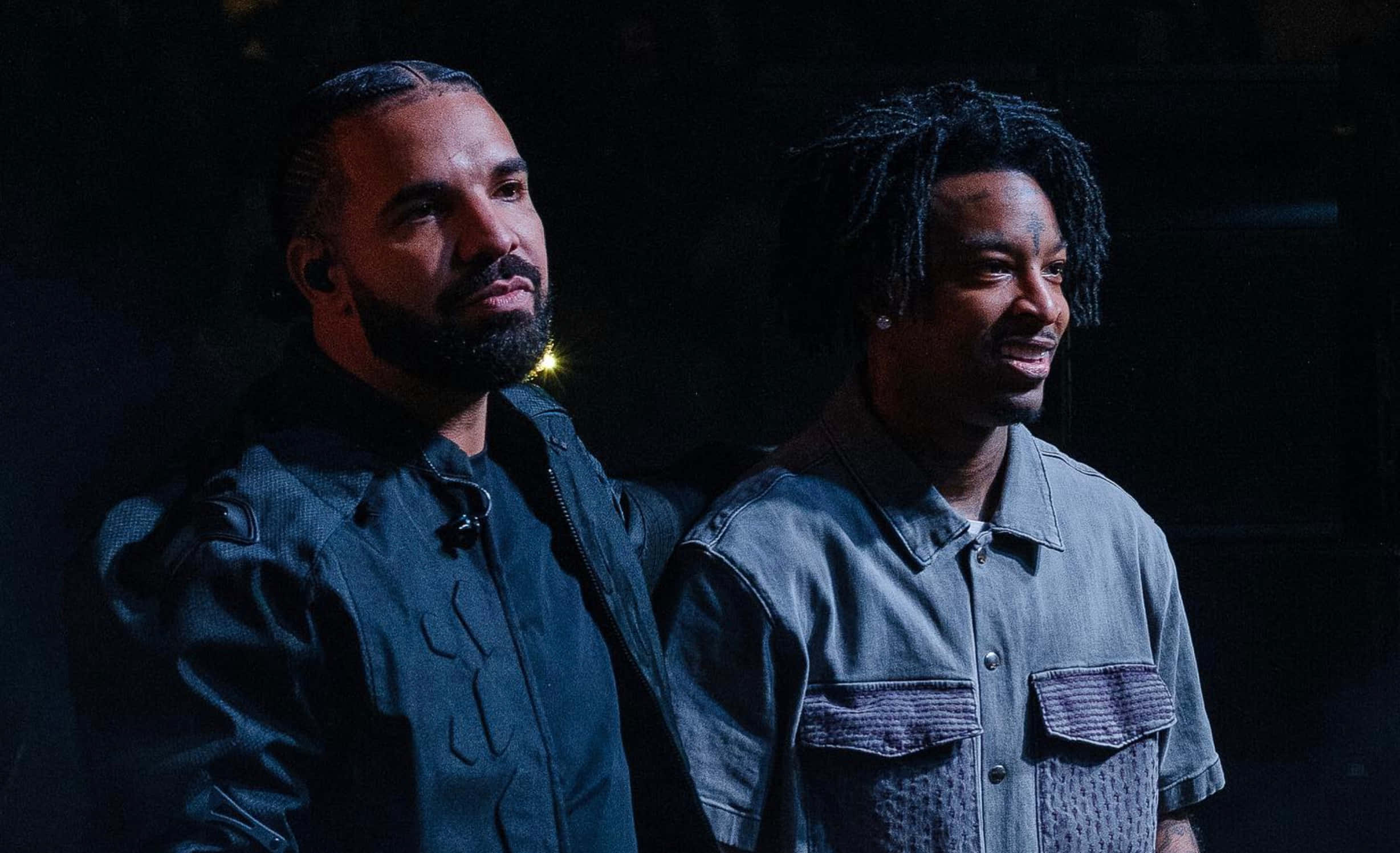 21 Savage 出席 MTV 音樂錄影帶大獎 壁紙 — 兩名身穿深色衣服的男子在黑色背景前，於戲劇性的舞台燈光下並肩站立。