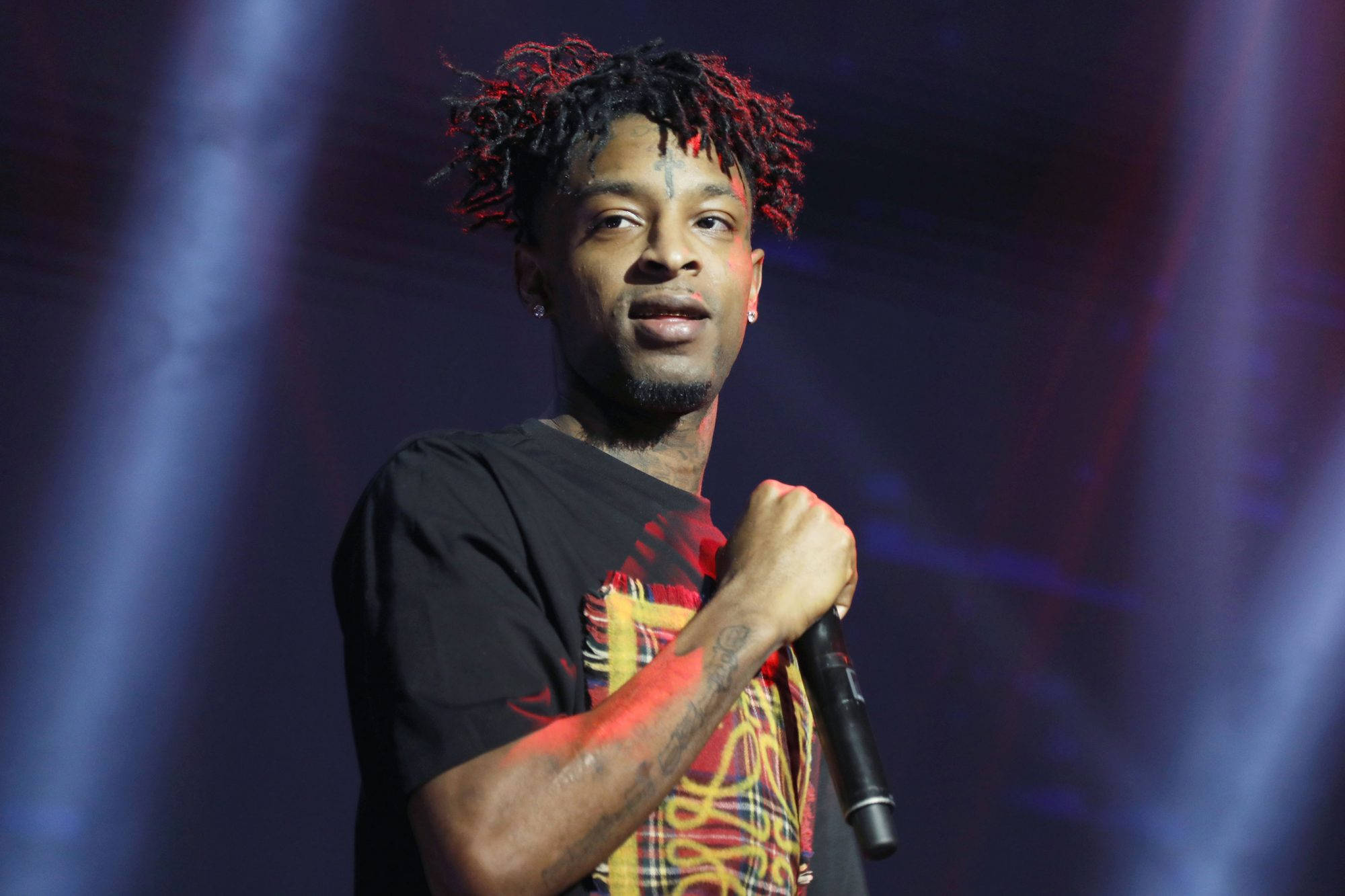 21 Savage 出席 Rolling Stone Live 活動 壁紙 — 一位男饒舌歌手留著紅色尖端的辮子，在戲劇性的藍色和紫色燈光下拿著麥克風在舞台上表演。