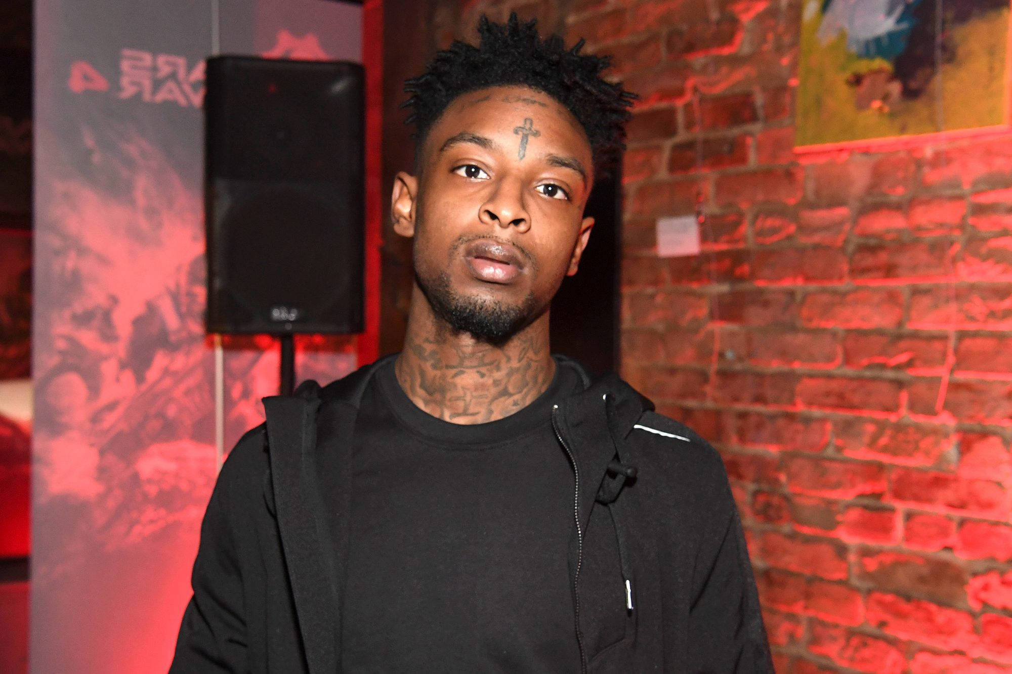 21 Savage 出席 Xbox 亞特蘭大活動 壁紙 — 一個額頭上有十字架紋身的男人，站在紅光照耀的磚牆前，穿著黑色連帽衫。