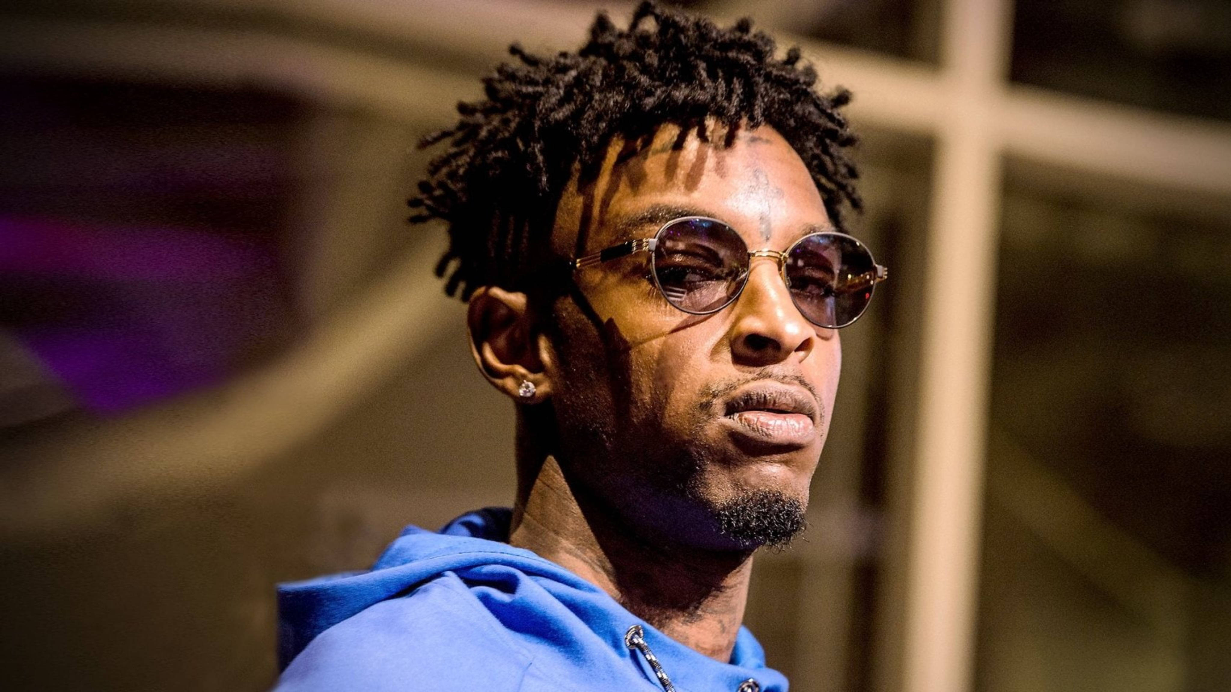 21 Savage 出席 YouTube Space 壁紙 — 一個捲髮、戴圓形太陽眼鏡的男人，在溫暖的夜間燈光下穿著藍色連帽衫。