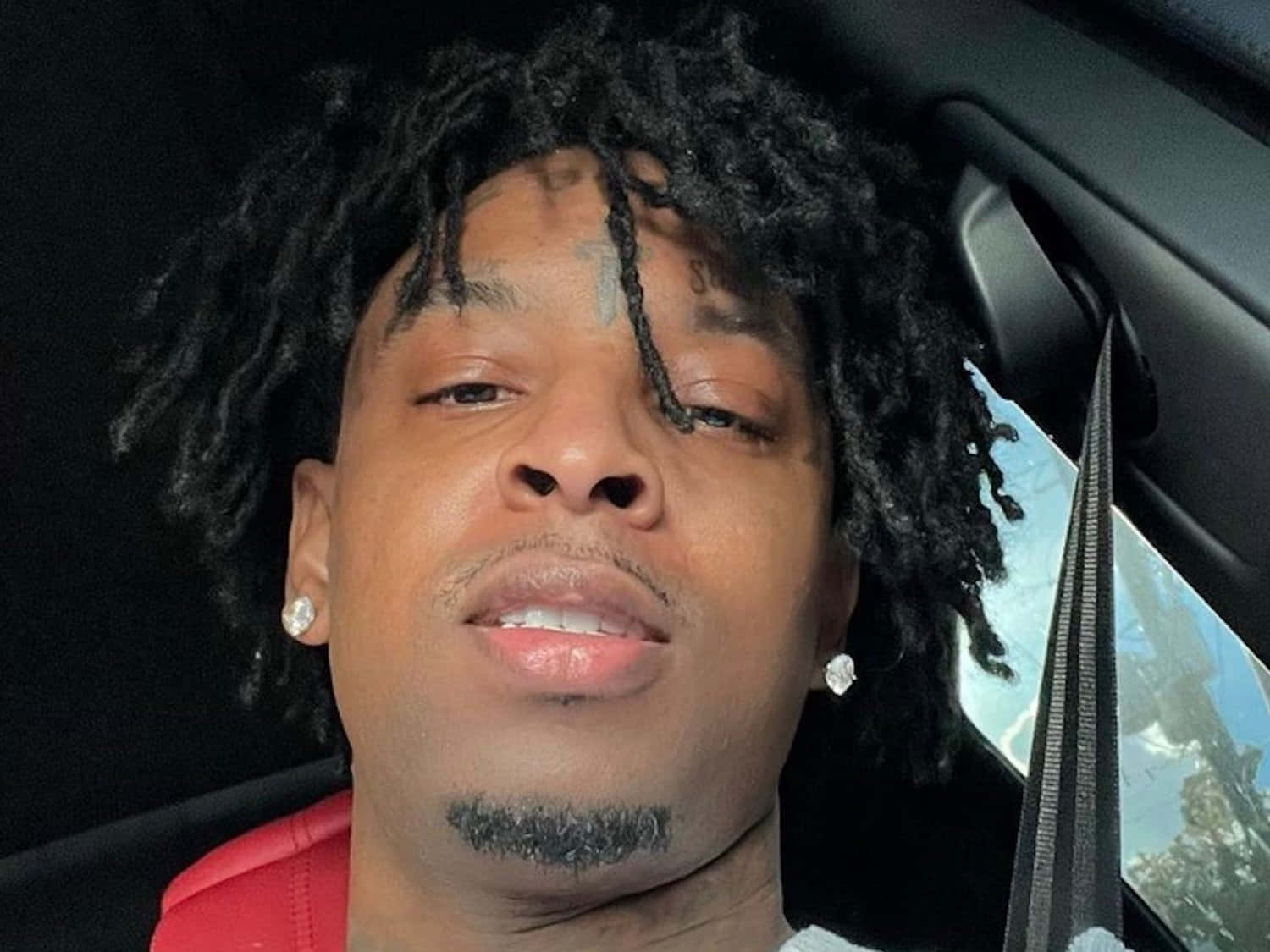 "21 Savage - 嘻哈傳奇" 壁紙 — 一名留著辮子頭、臉上有紋身的男子，身穿紅色襯衫坐在車內的近距離特寫。