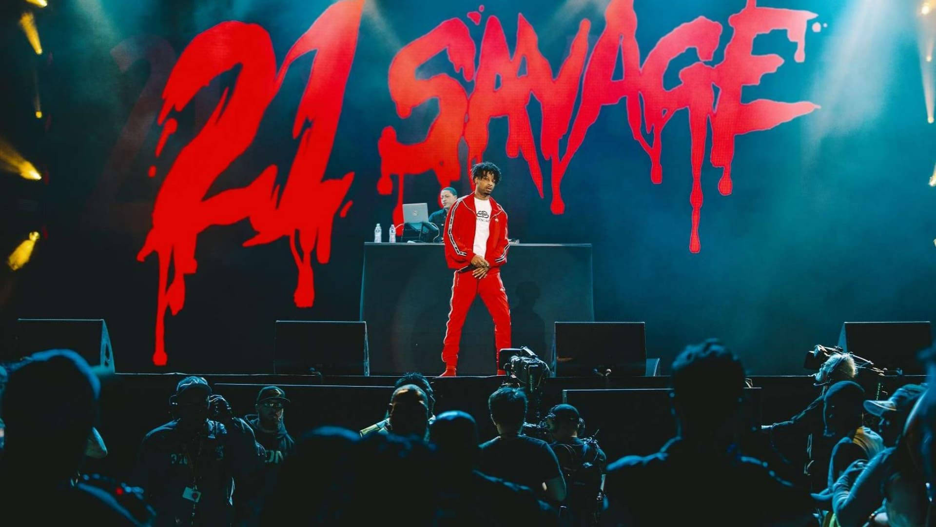 21 Savage 在夢幻村音樂節演出 壁紙 — 一位身穿紅色運動服的饒舌歌手在藍色燈光下的黑暗舞台上表演，背景是滴落的紅色「21 SAVAGE」。