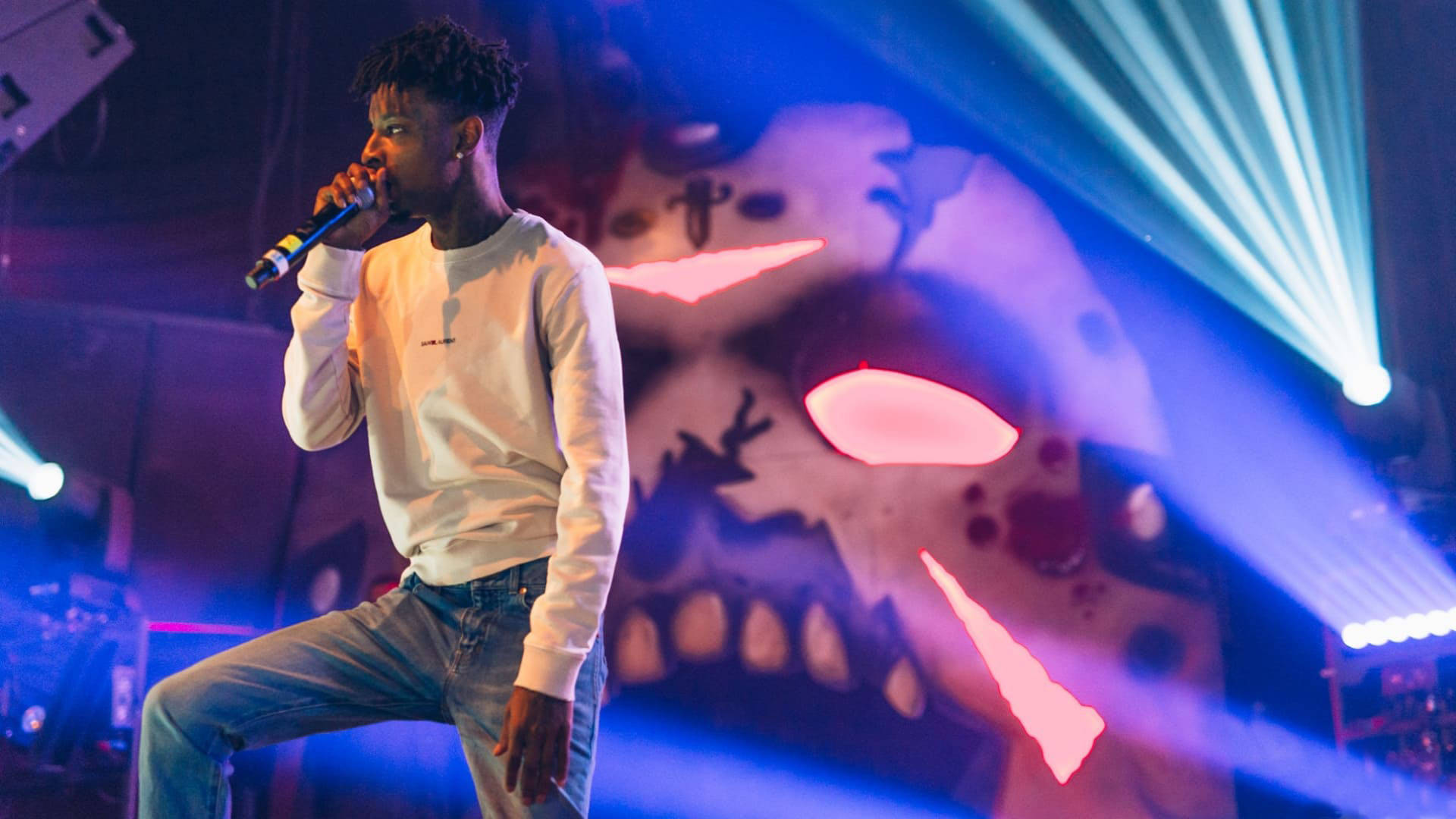 21 Savage 在奧格登劇院演出 壁紙 — 一位男饒舌歌手在藍色燈光下表演，背景是一個巨大的發光骷髏頭。