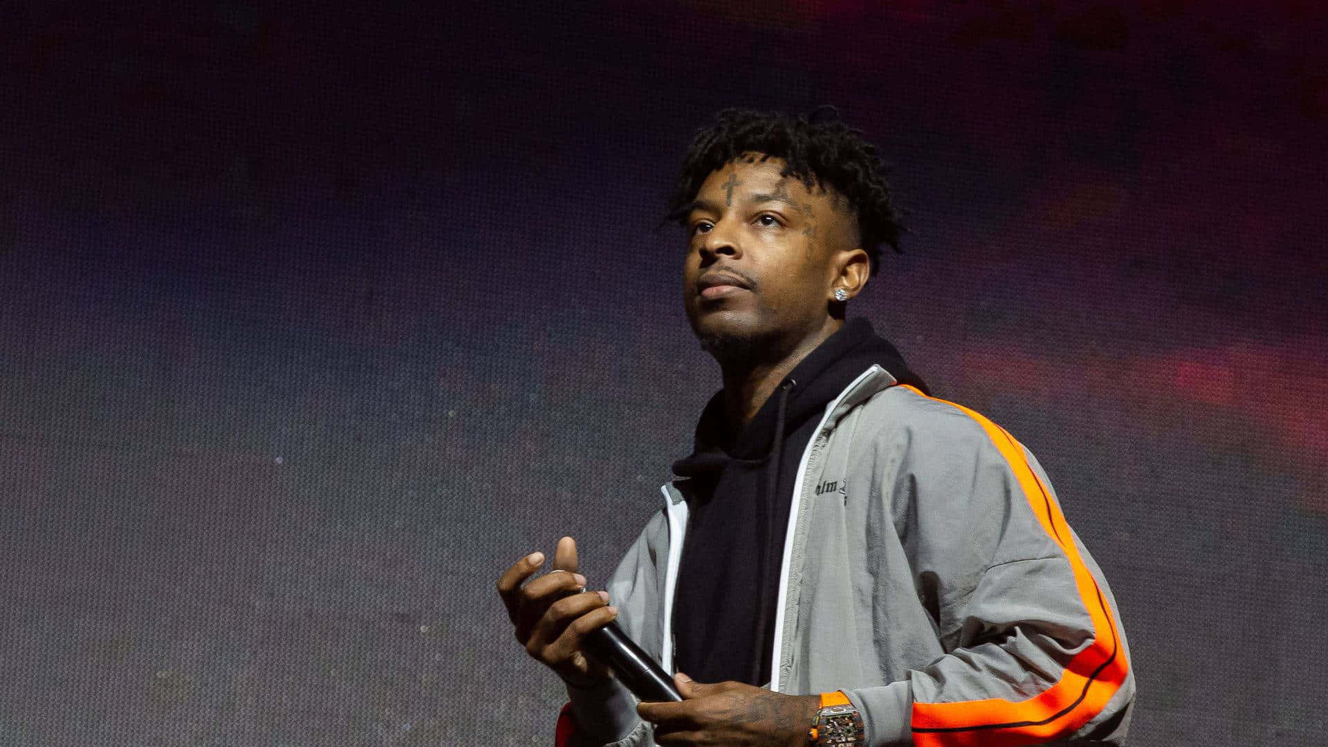 21 Savage 在攝影棚拍攝 壁紙 — 一位留著辮子的男性表演者在舞台上，在戲劇性的紫色和紅色燈光下握著麥克風。