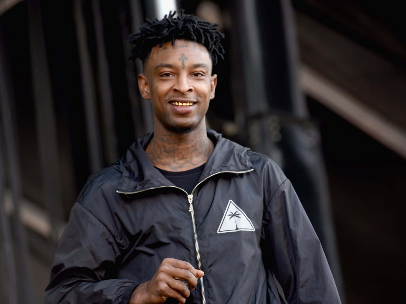 21 Savage 在格萊美頒獎典禮後的演出 壁紙 — 一個微笑的男人，留著辮子頭和紋身，穿著黑色外套，上面有白色棕櫚樹標誌，背景是黑暗的城市。