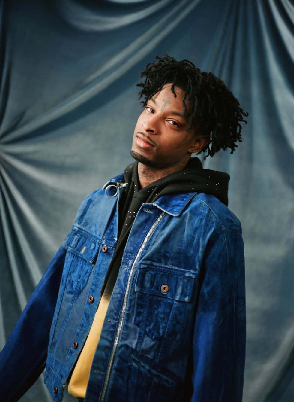 21 Savage 在演唱會表演 壁紙 — 一個有捲髮和面部紋身的男人，在藍色垂簾背景前，穿著藍色牛仔外套，內搭黑色連帽衫。