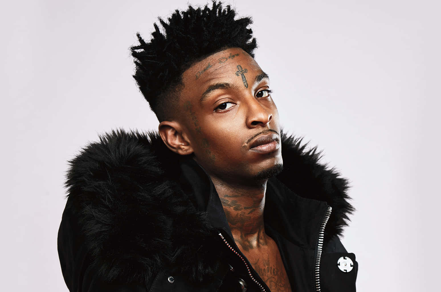 21 Savage 在現場舞台演出 壁紙 — 一個有黑色尖刺頭髮和面部紋身的男人，在純白色背景前穿著黑色毛領夾克。