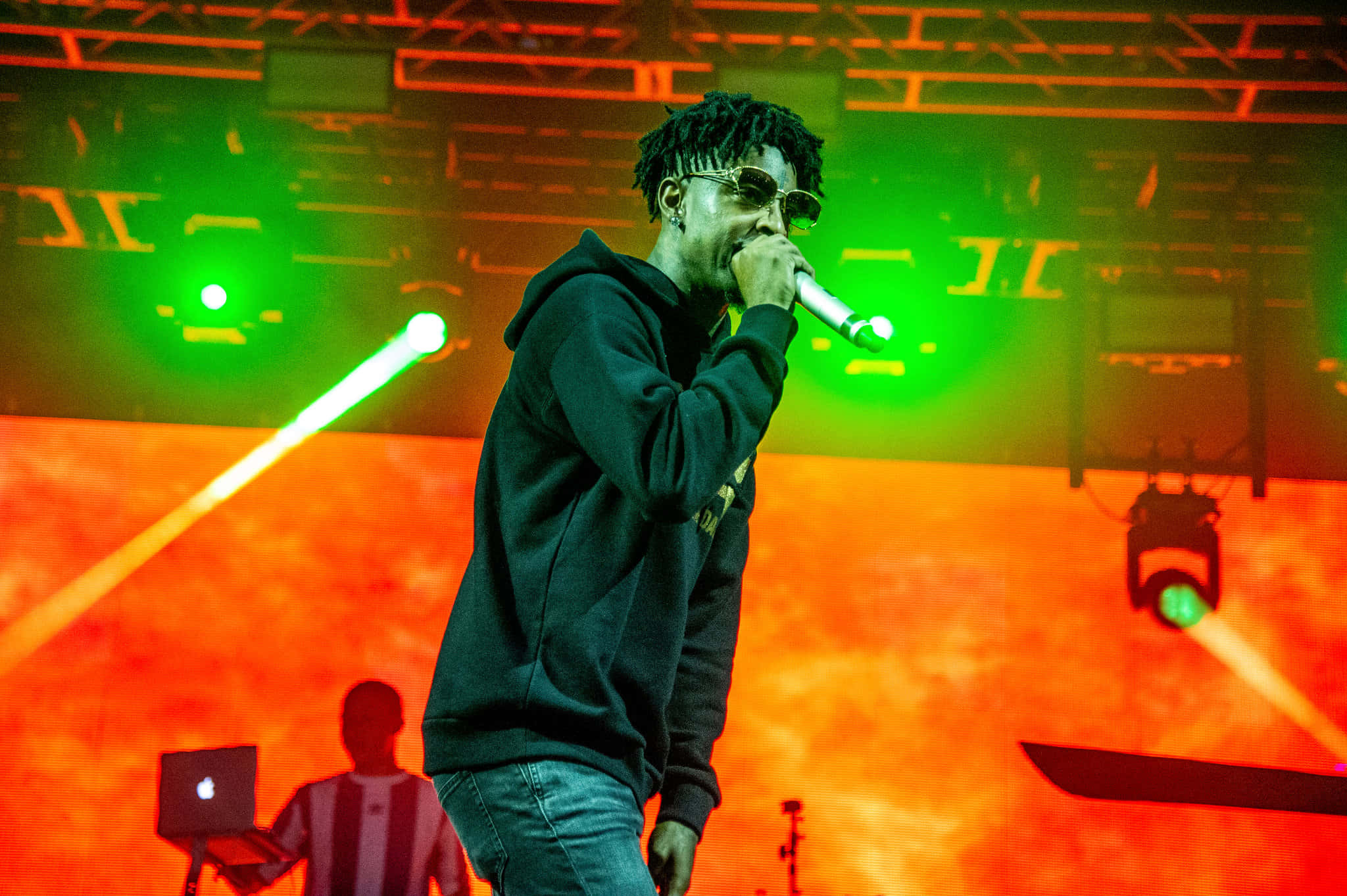 21 Savage 在科切拉音樂節演出 壁紙 — 一位戴著太陽眼鏡和連帽衫的饒舌歌手在舞台上表演，背景是鮮豔的綠色和橙色燈光，還有一位筆記型電腦技術人員。