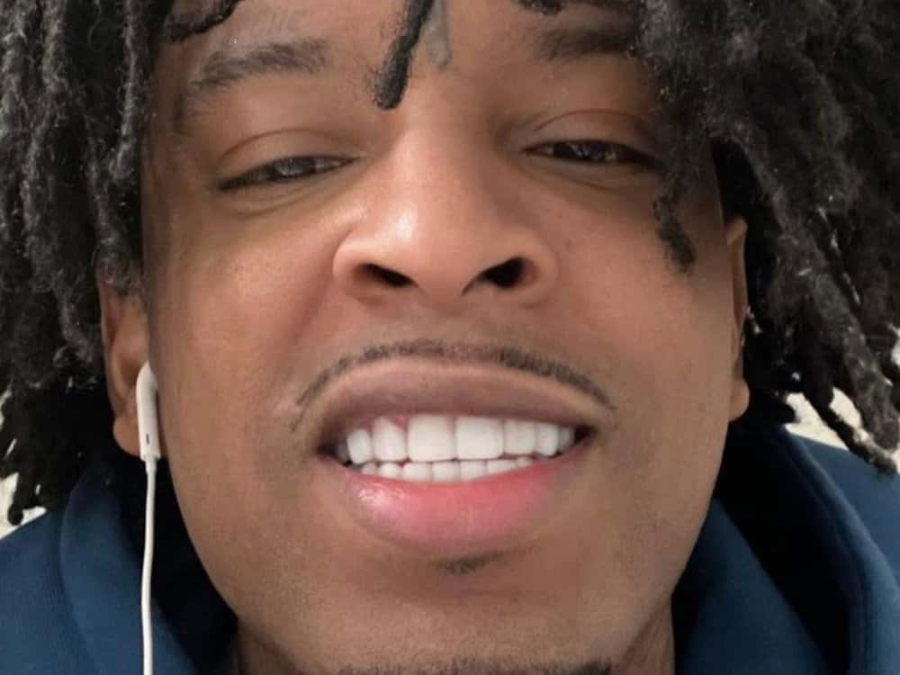 21 Savage 在錄音室發表宣言 壁紙 — 特寫一個微笑的男人，深色辮子，白色耳塞，穿著藍色連帽衫。