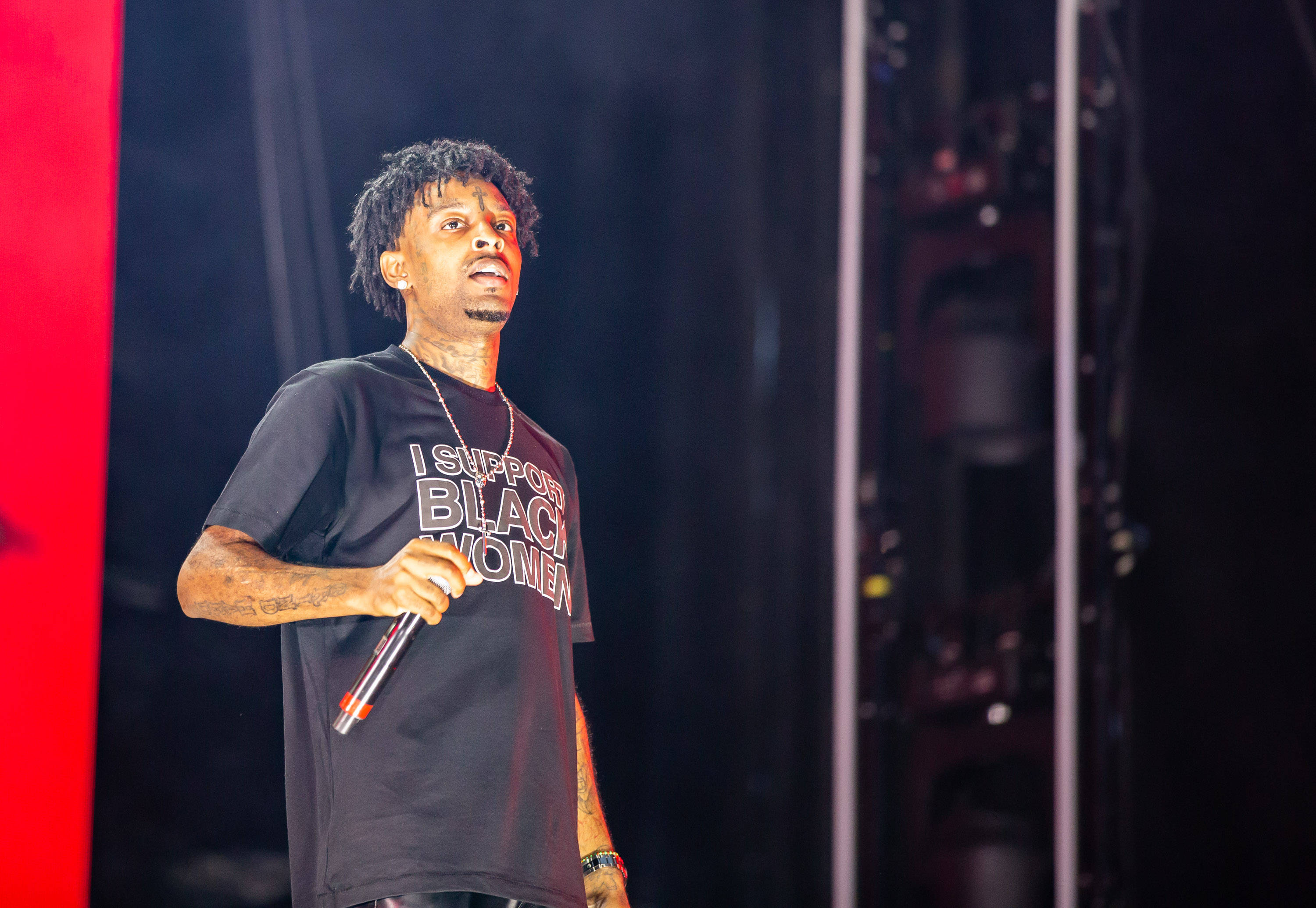 21 Savage 在音樂中城音樂節演出 壁紙 — 一位帶有紋身、身穿黑色T恤（上面寫著「我支持黑人女性」）的男性表演者，在燈光昏暗、紅色燈光的舞台上對著麥克風唱歌。