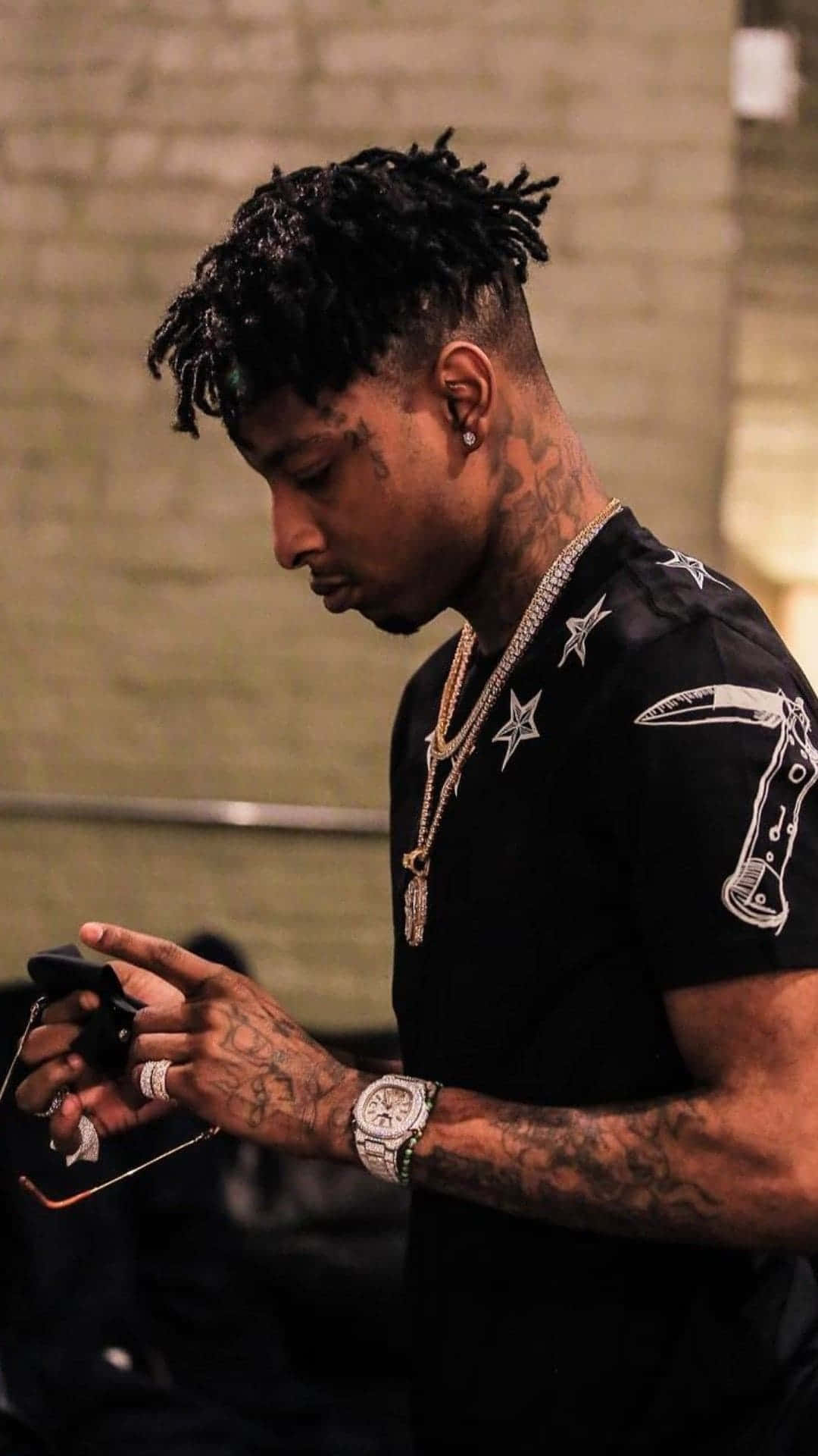 21 Savage 在麥克風前 壁紙 — 一名留著雷鬼辮、有紋身的男子，穿著黑色星星印花襯衫和鑽石鏈條，低頭看著手機。