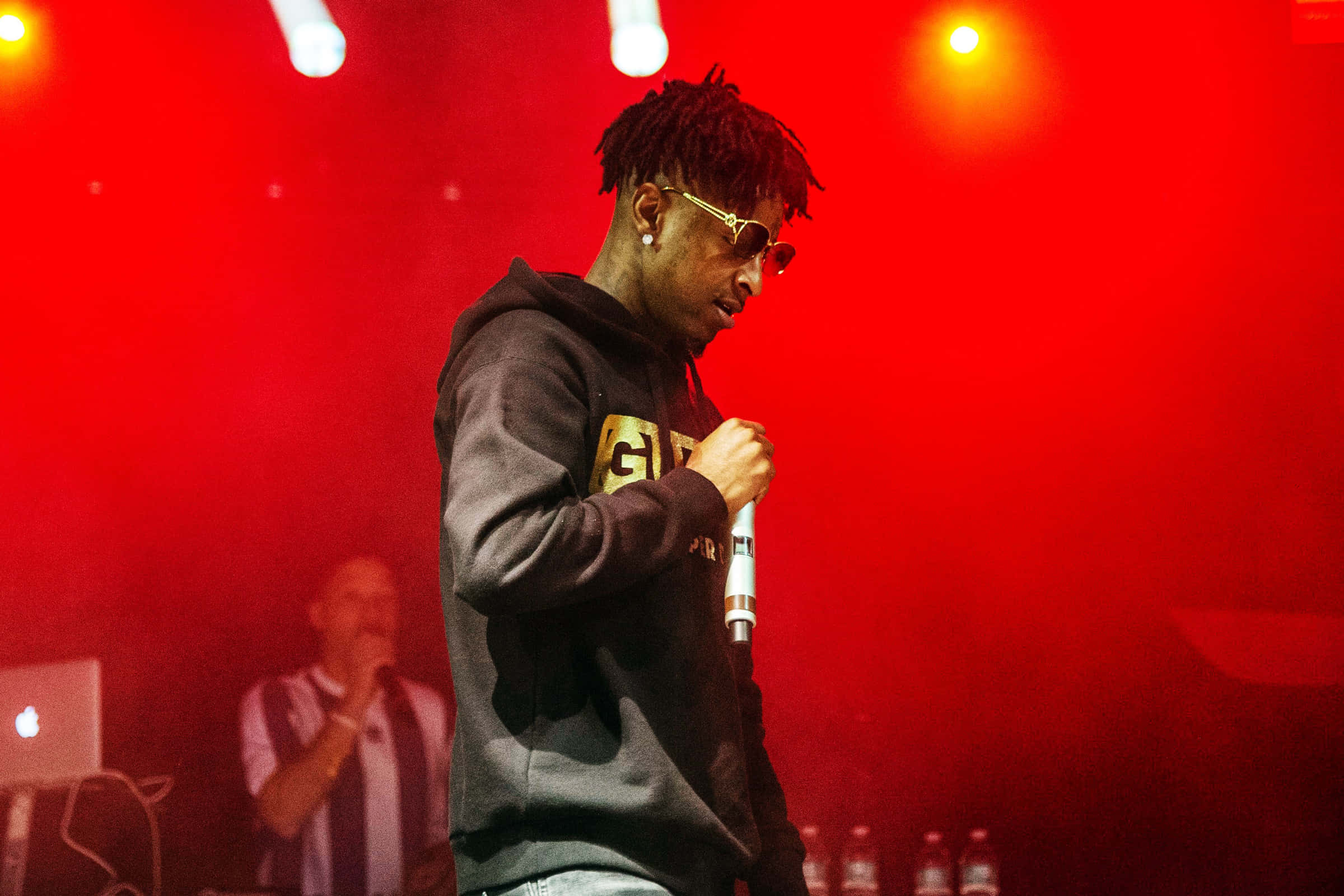 21 Savage 展現「Savage Mode」標誌 壁紙 — 一位留著辮子頭、戴金邊眼鏡的饒舌歌手在強烈紅光下拿著麥克風在舞台上表演。