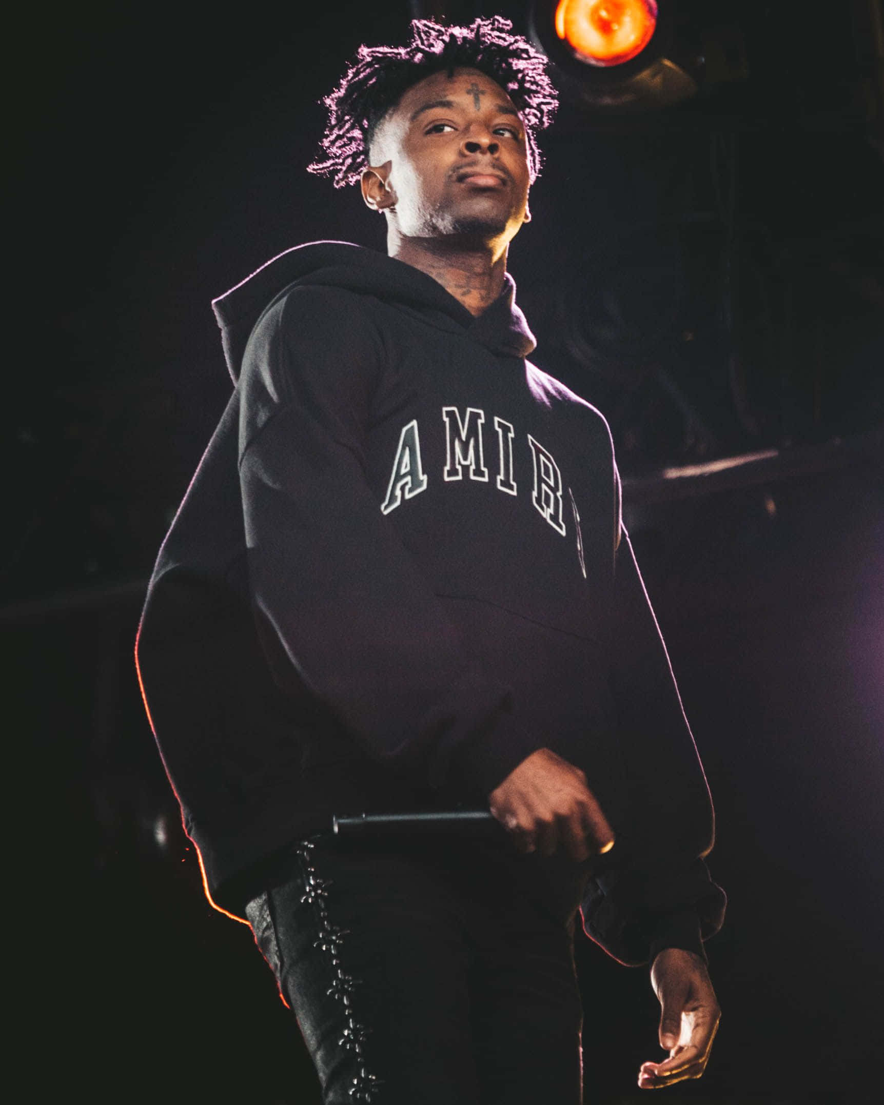 21 Savage 展現饒舌實力 壁紙 — 一位捲髮、身穿連帽衫的男性表演者站在燈光昏暗的舞台上，在溫暖的聚光燈下握著麥克風。