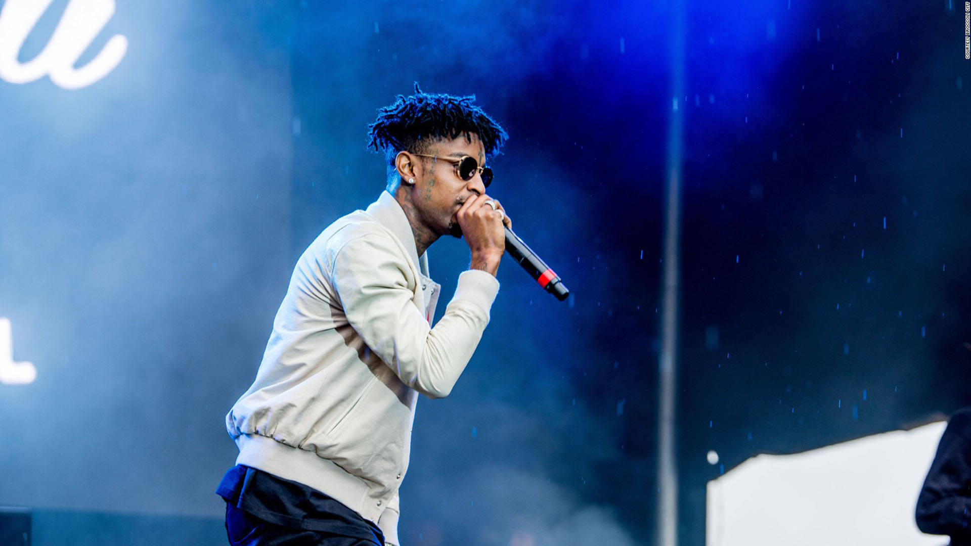 21 Savage 布羅ccoli 城市音樂節 壁紙 — 一位梳著辮子頭、戴著太陽眼鏡的饒舌歌手在藍色舞台燈光下對著麥克風表演，周圍下著雨。