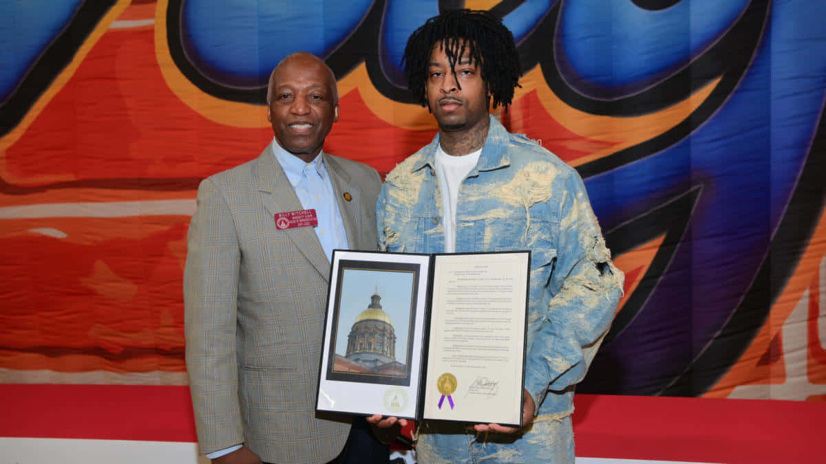 21 Savage - 捍衛陷阱音樂之王 壁紙 — 兩名男子微笑著手持裱框證書和肖像，背景是充滿活力的塗鴉風格。