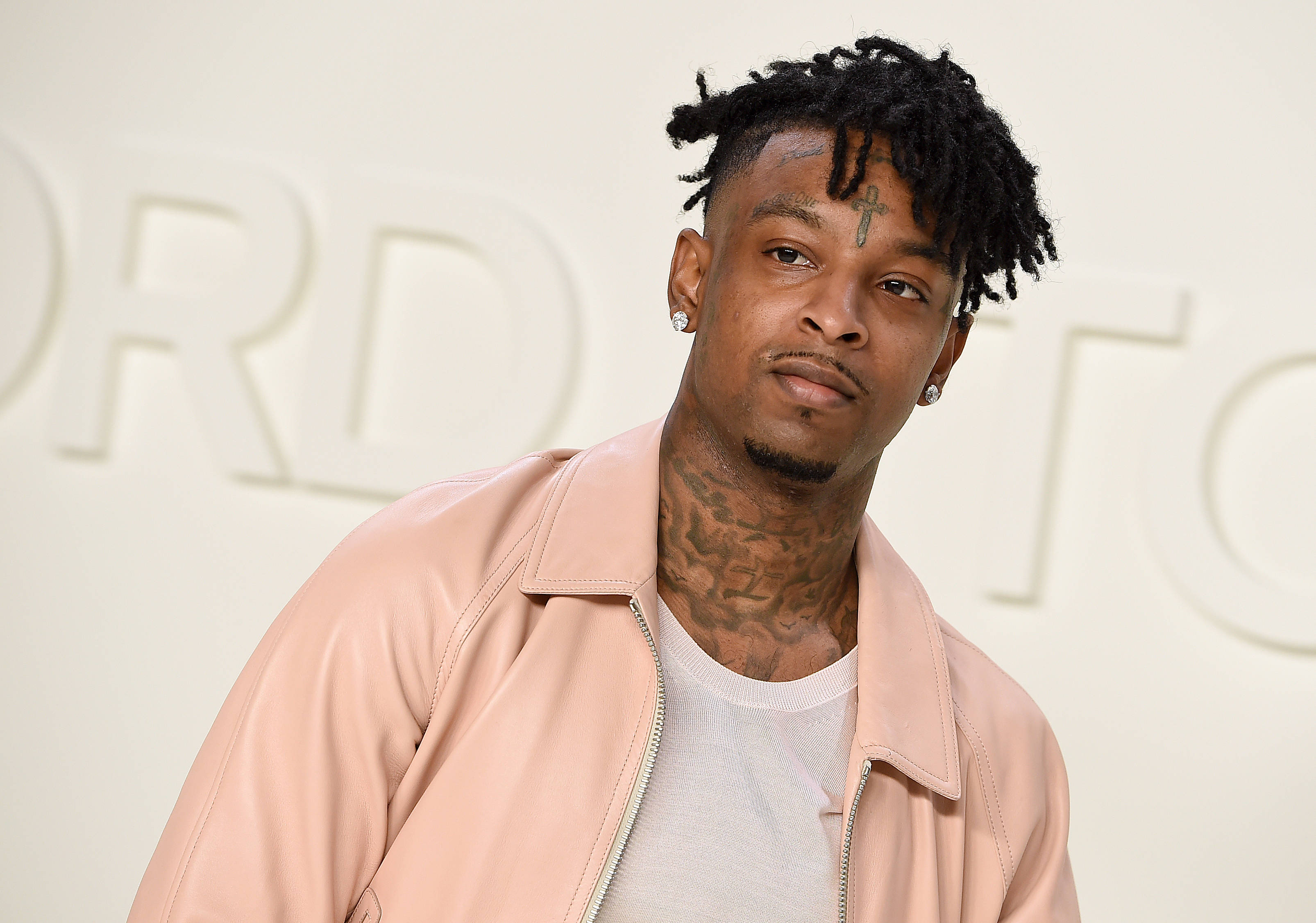 21 Savage 湯姆·福特 AW20 時裝秀 壁紙 — 一名留著辮子頭、頸部有紋身的男子，在簡約白色背景（帶有模糊字樣）前穿著粉紅色皮褸。