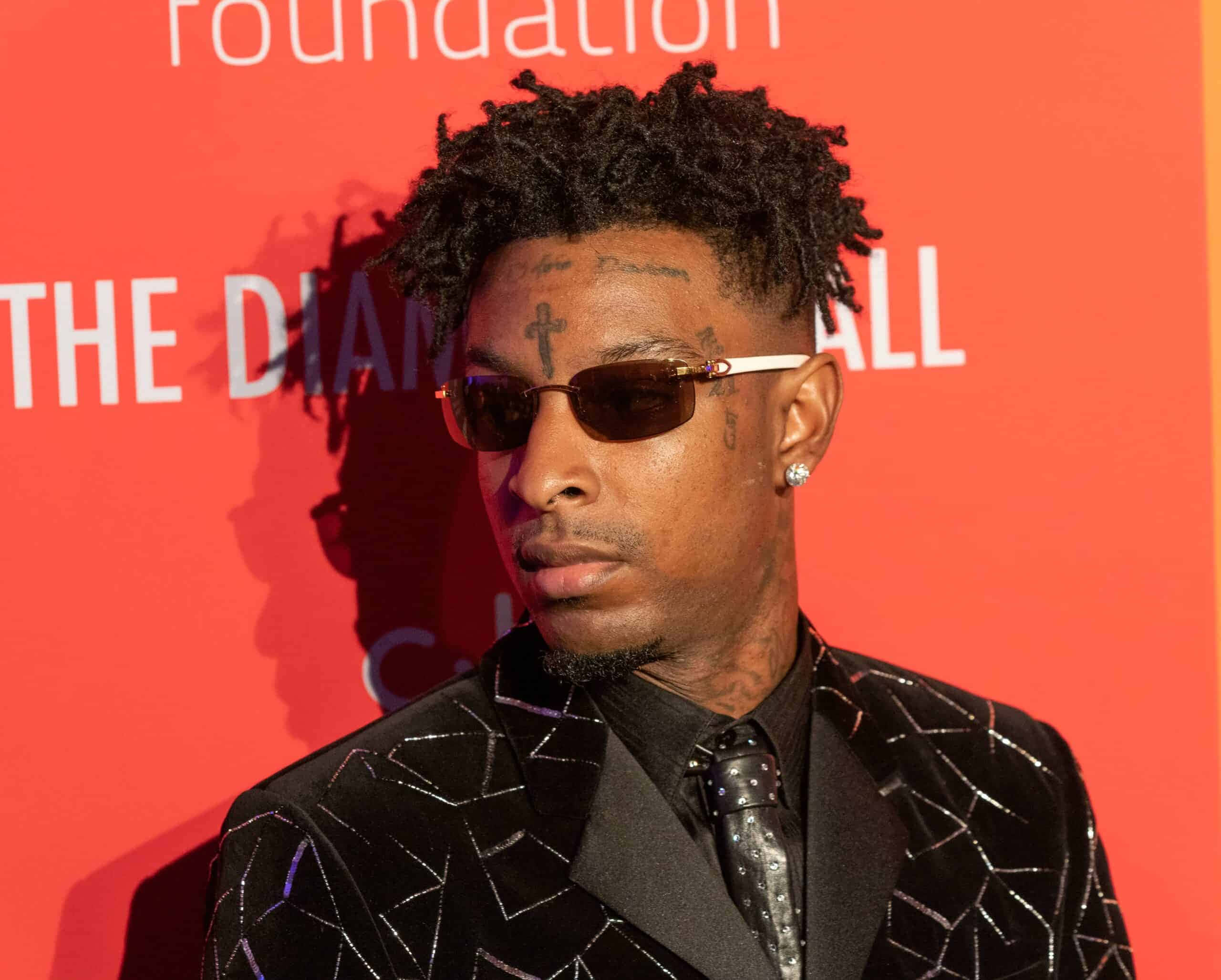 21 Savage 演唱會現場演出 壁紙 — 一名留著辮子頭和紋身的男子，戴著太陽眼鏡，穿著閃亮的黑色西裝，背景是紅色活動背景。