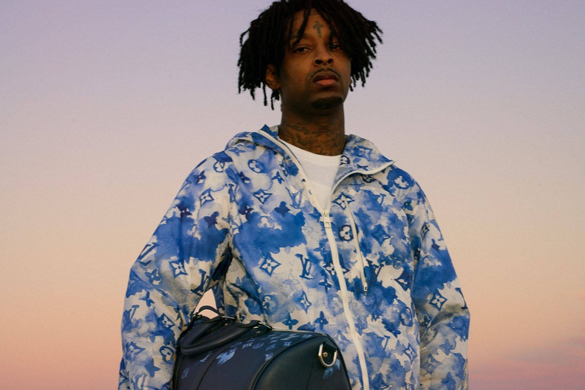 21 Savage 穿 Louis Vuitton 夾克 壁紙 — 一個有辮子頭和紋身的男人，穿著藍色圖案連帽衫，背景是柔和的粉紅色和紫色日落天空。
