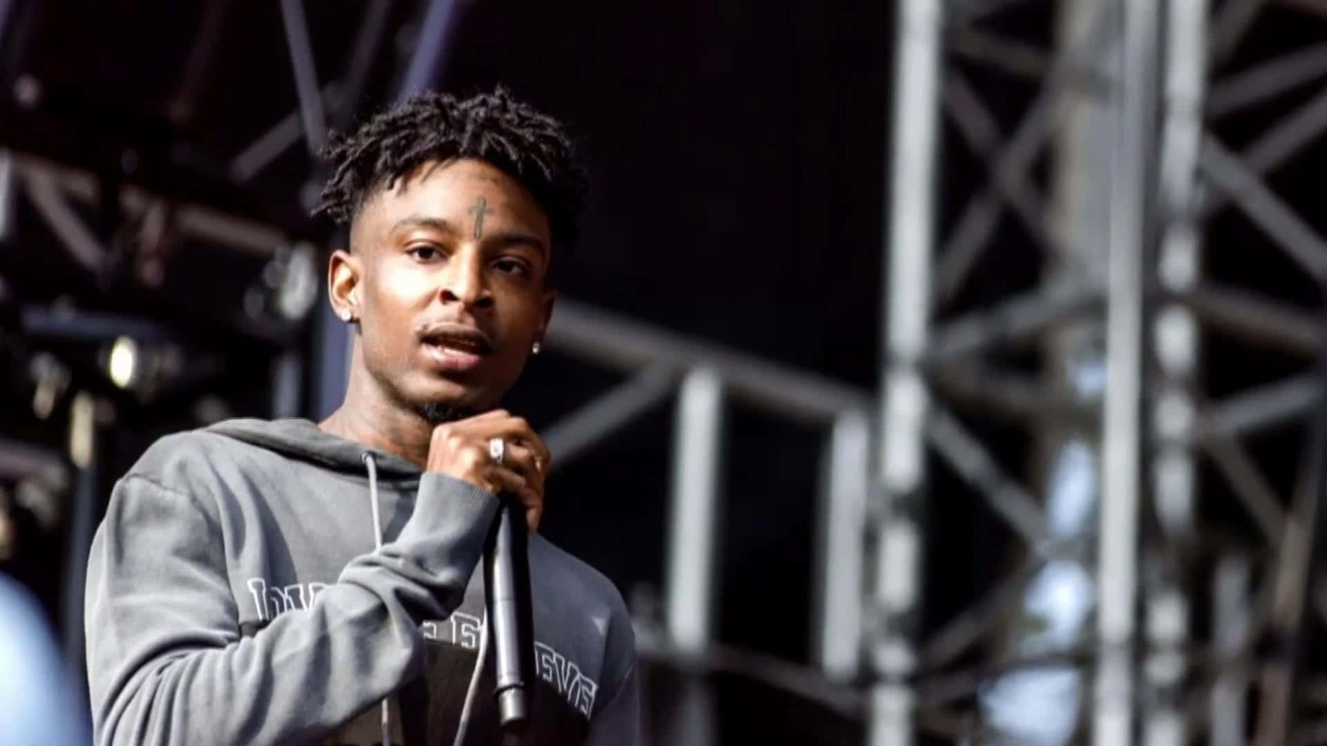 21 Savage 穿白色夾克拍攝 壁紙 — 一位額頭上有十字紋身的年輕男饒舌歌手在舞台燈光下熱情地對著麥克風表演。
