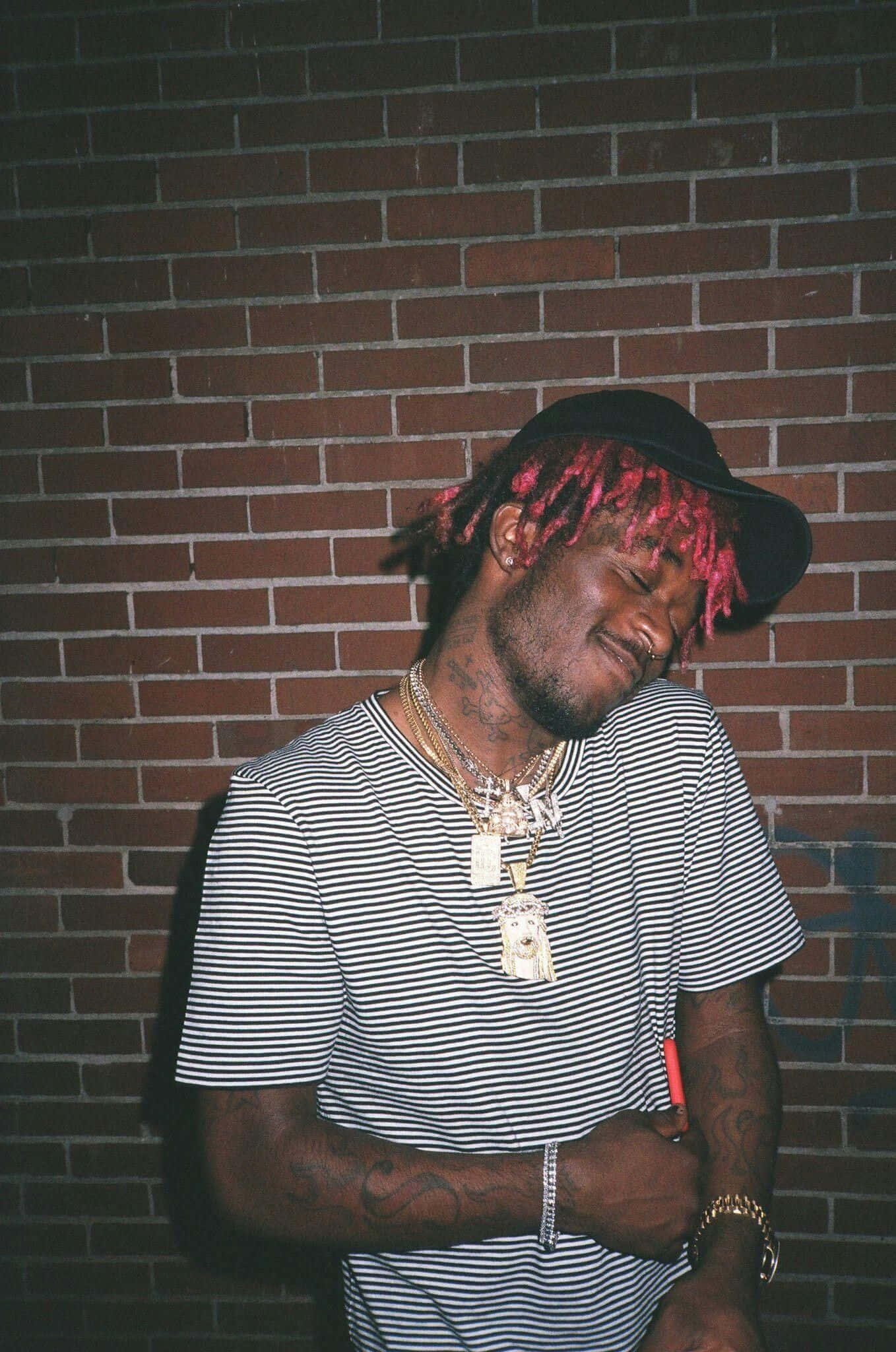 21 Savage 站在「Savage Mode」混音帶背景前 壁紙 — 一個留著粉紅色辮子、戴著金鏈的男人在晚上靠著磚牆，閉著眼睛，姿勢放鬆。