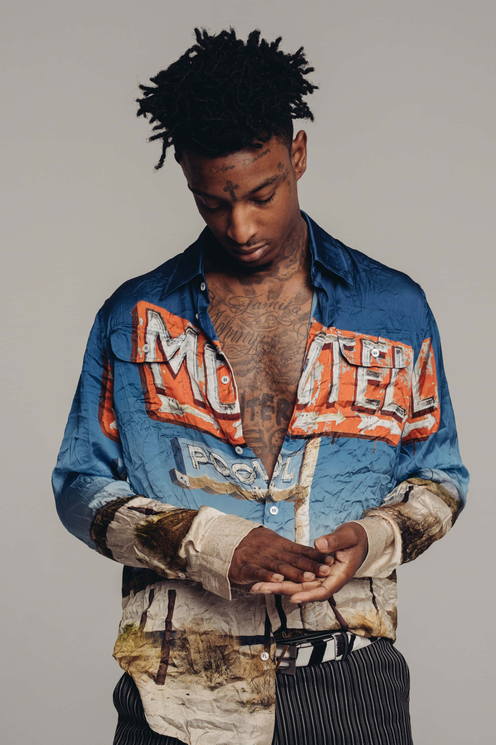 21 Savage 站在標誌性 XO 品牌標誌前 壁紙 — 一位留著辮子頭和紋身的年輕男子，穿著一件帶有橙色字樣的破舊藍色襯衫，低頭看著自己的手。