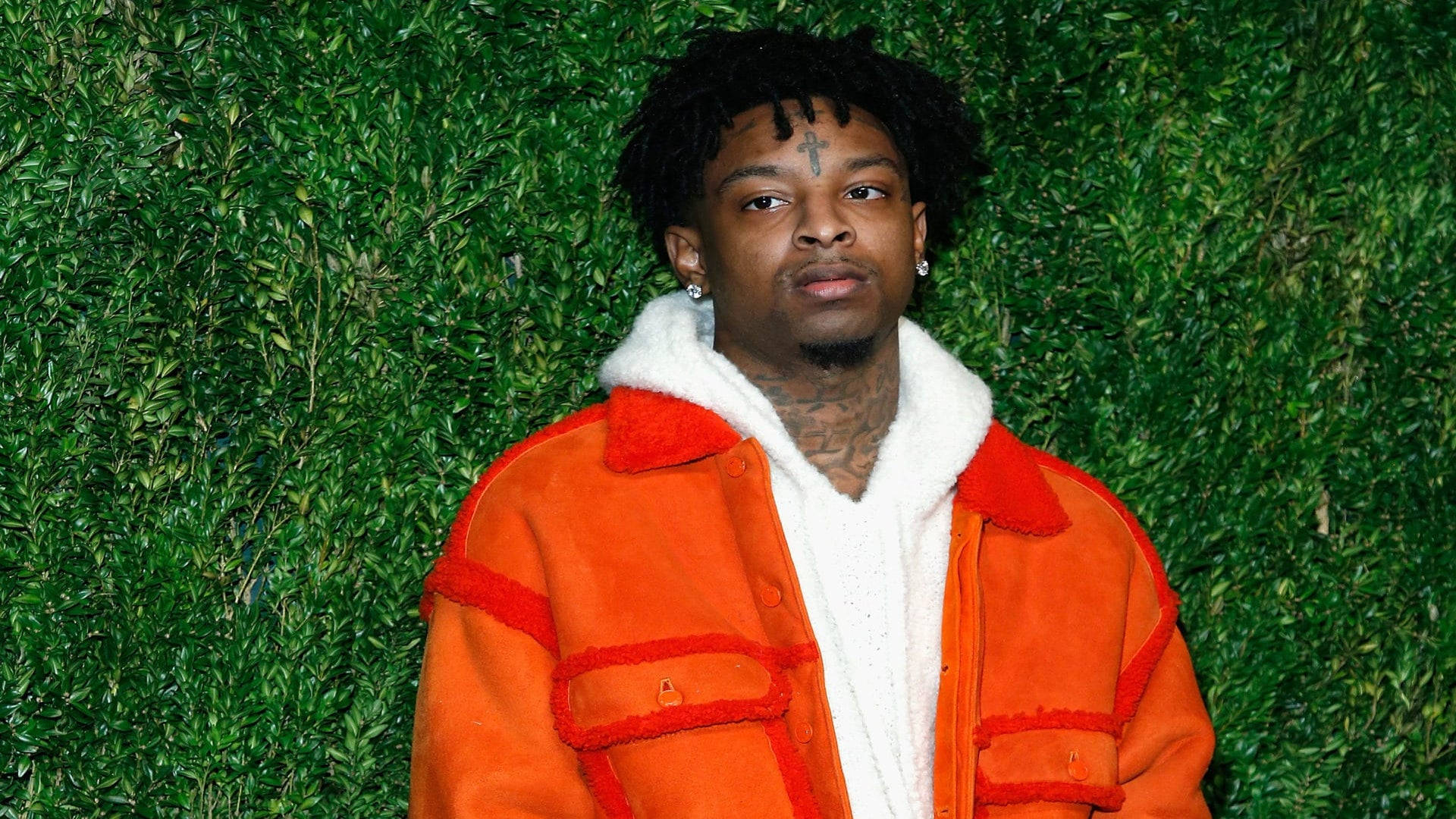 21 Savage 與 Vogue 時尚基金會 壁紙 — 一名留著辮子頭、有十字架紋身的男子，身穿白色毛領的亮橙色外套，凝視前方，背景是茂密的綠色樹籬牆。